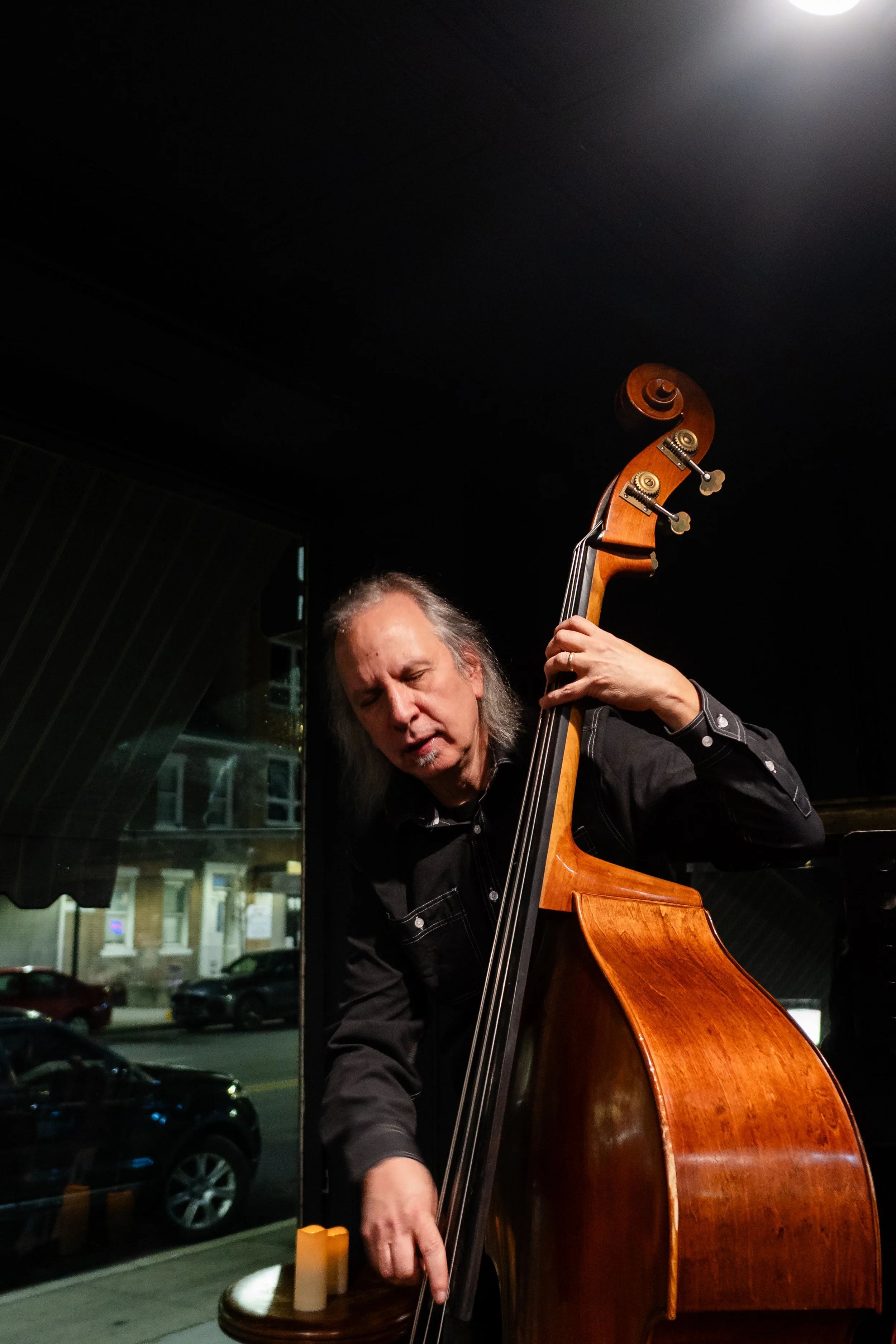 A man playing a double bass in a dark indoor setting with a street view visible through a large window