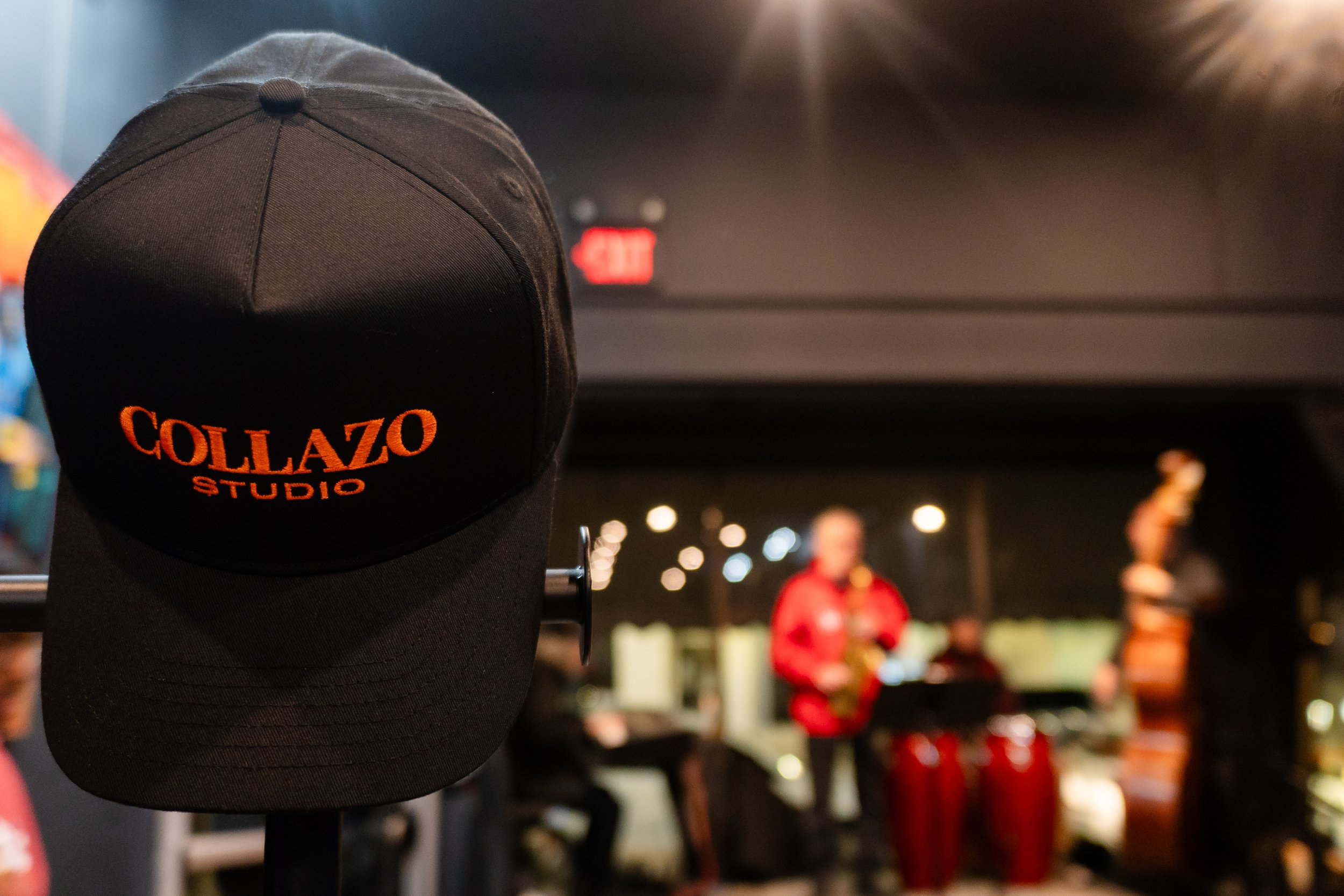 Black Cap with orange embroidery reading "COLLAZO STUDIO" hanging in a room with a blurred background of people, including one person in red, playing musical instruments.