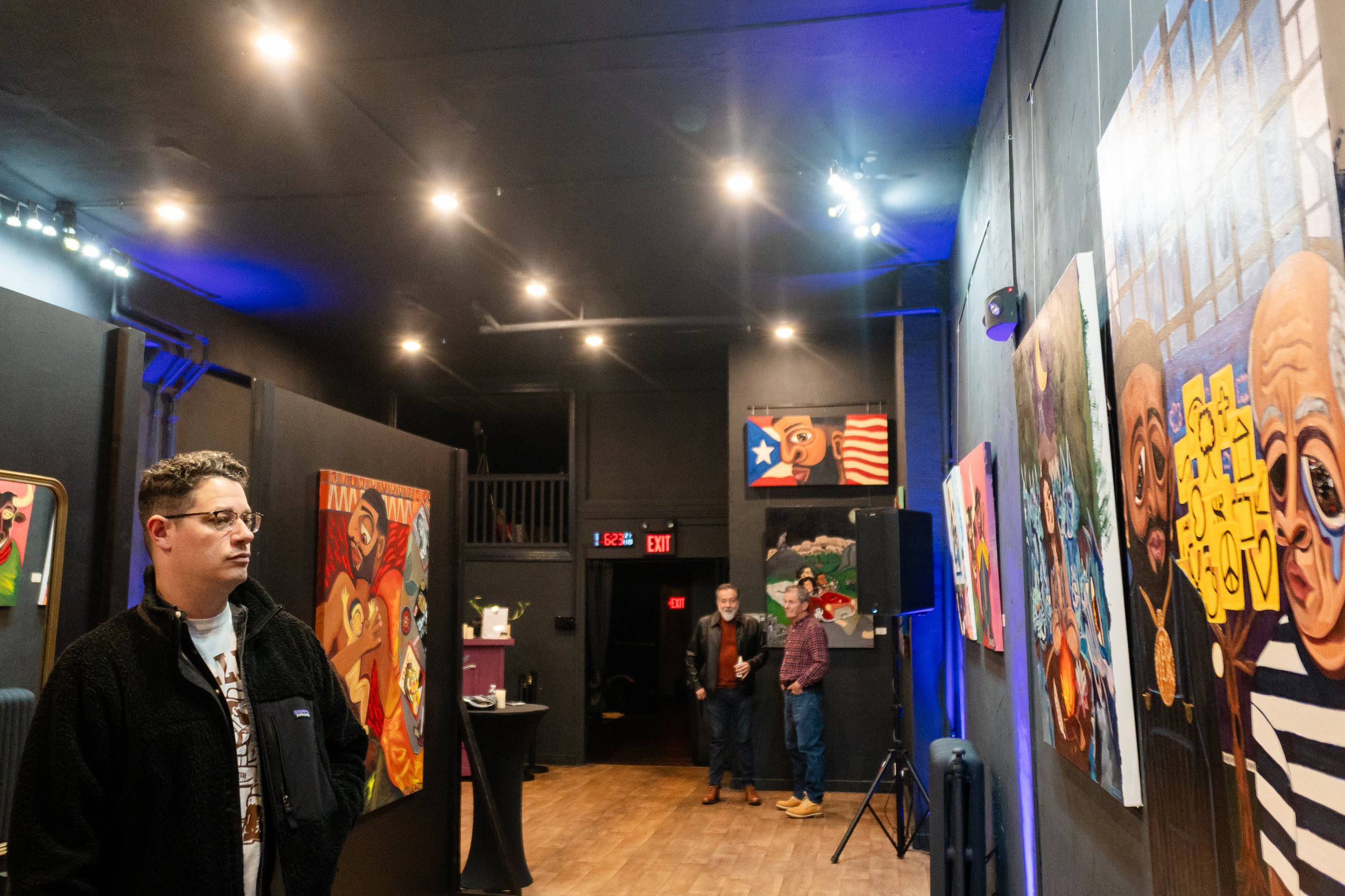 An art gallery with vibrant paintings on the walls and three men standing and conversing in the background, while one man in the foreground looks at the artwork.