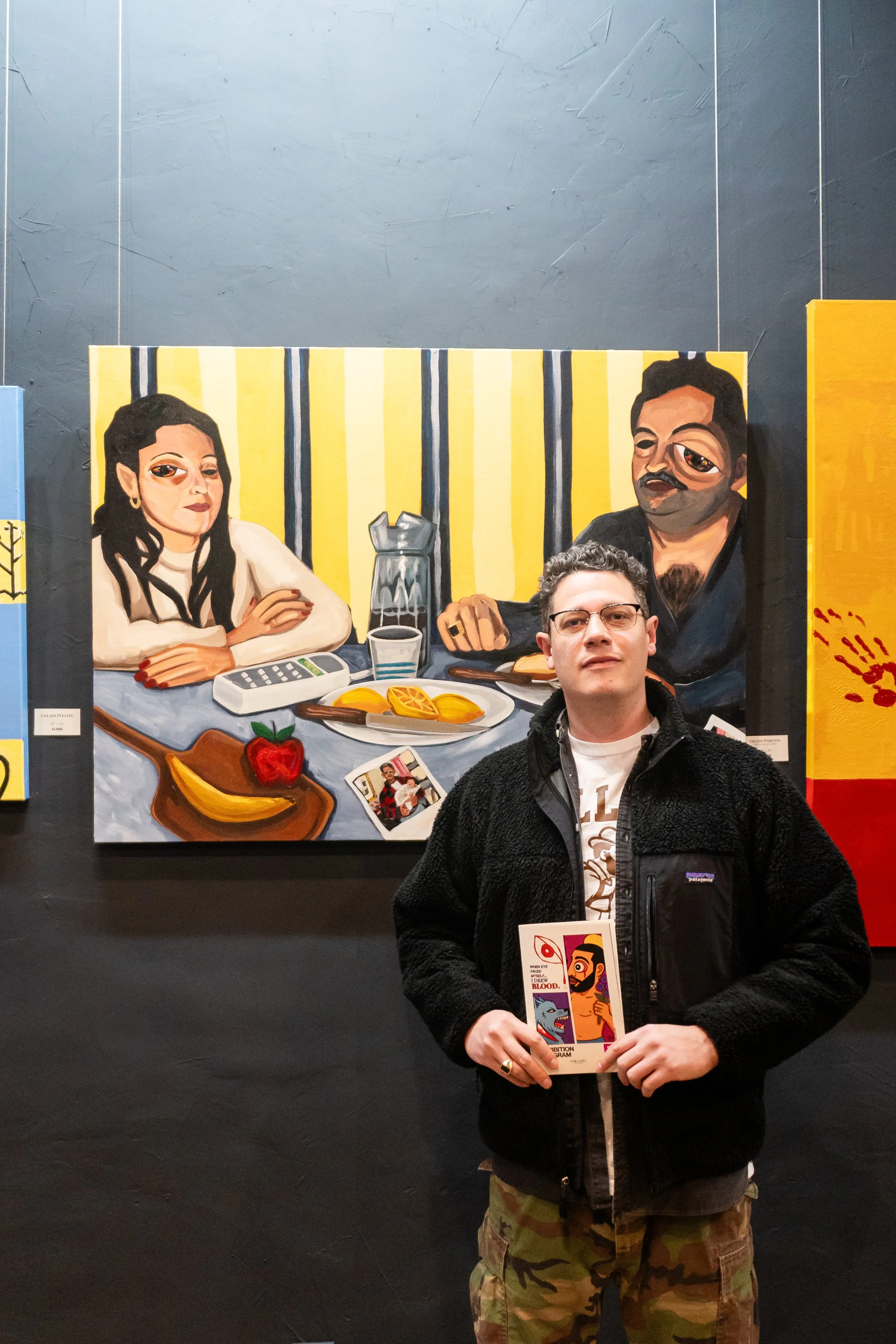 A man standing in front of a colorful painting in an art gallery, holding a brochure or pamphlet.