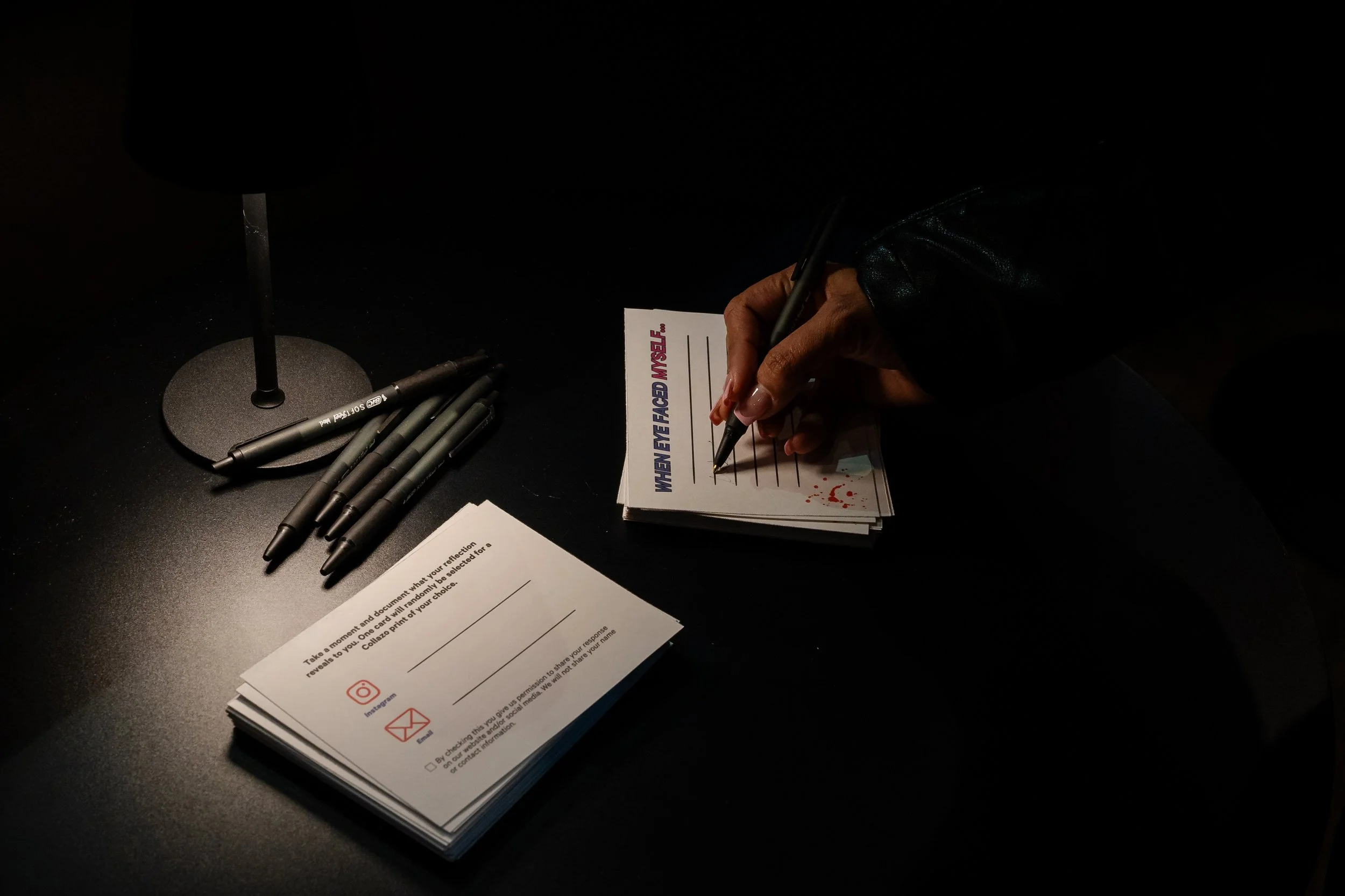 Person writing on a stack of postcards with a black pen, several black pens on the table, and a dark background.