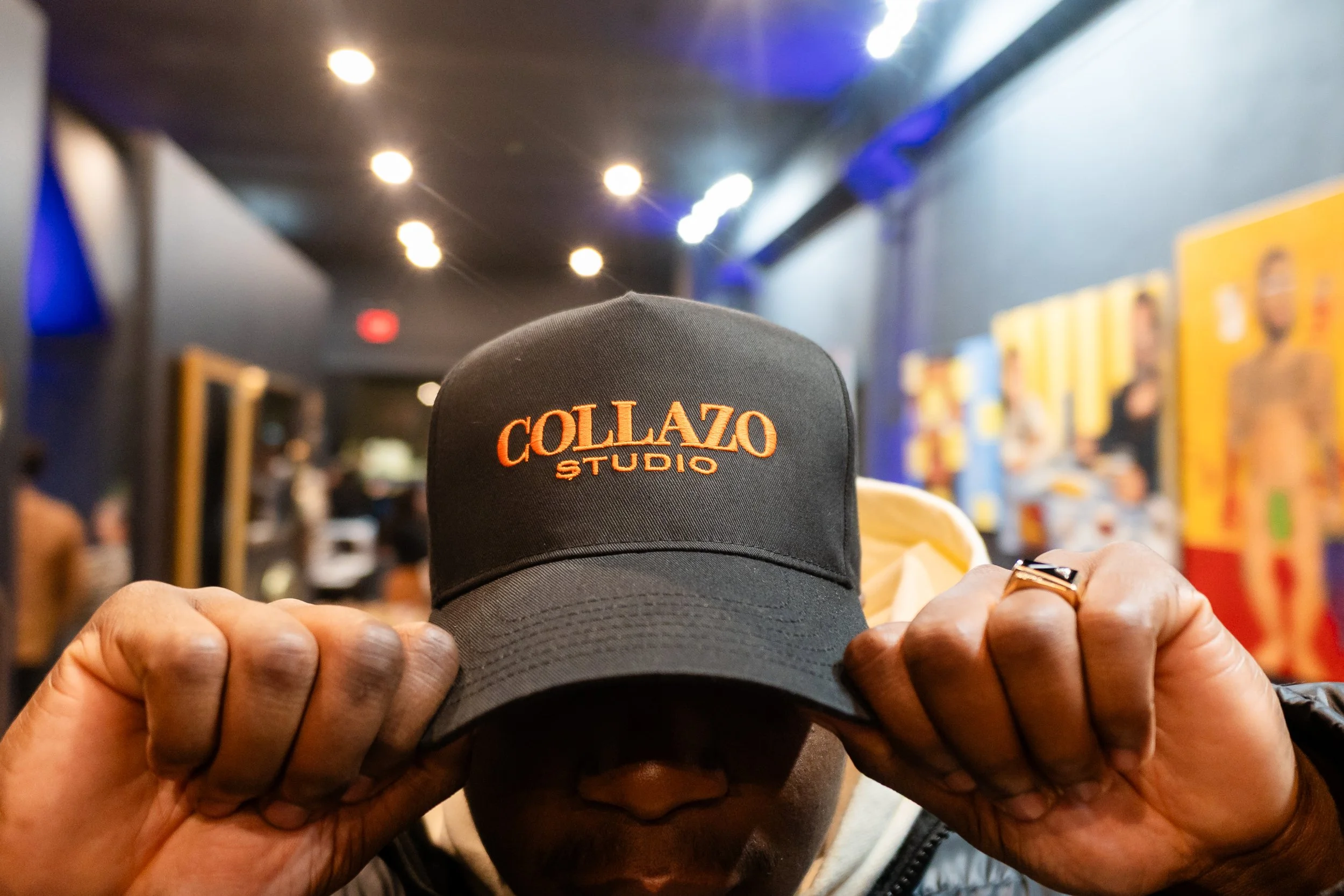 Person holding a black baseball cap with orange embroidered text 'COLLAZO STUDIO' in an art gallery or exhibition space.