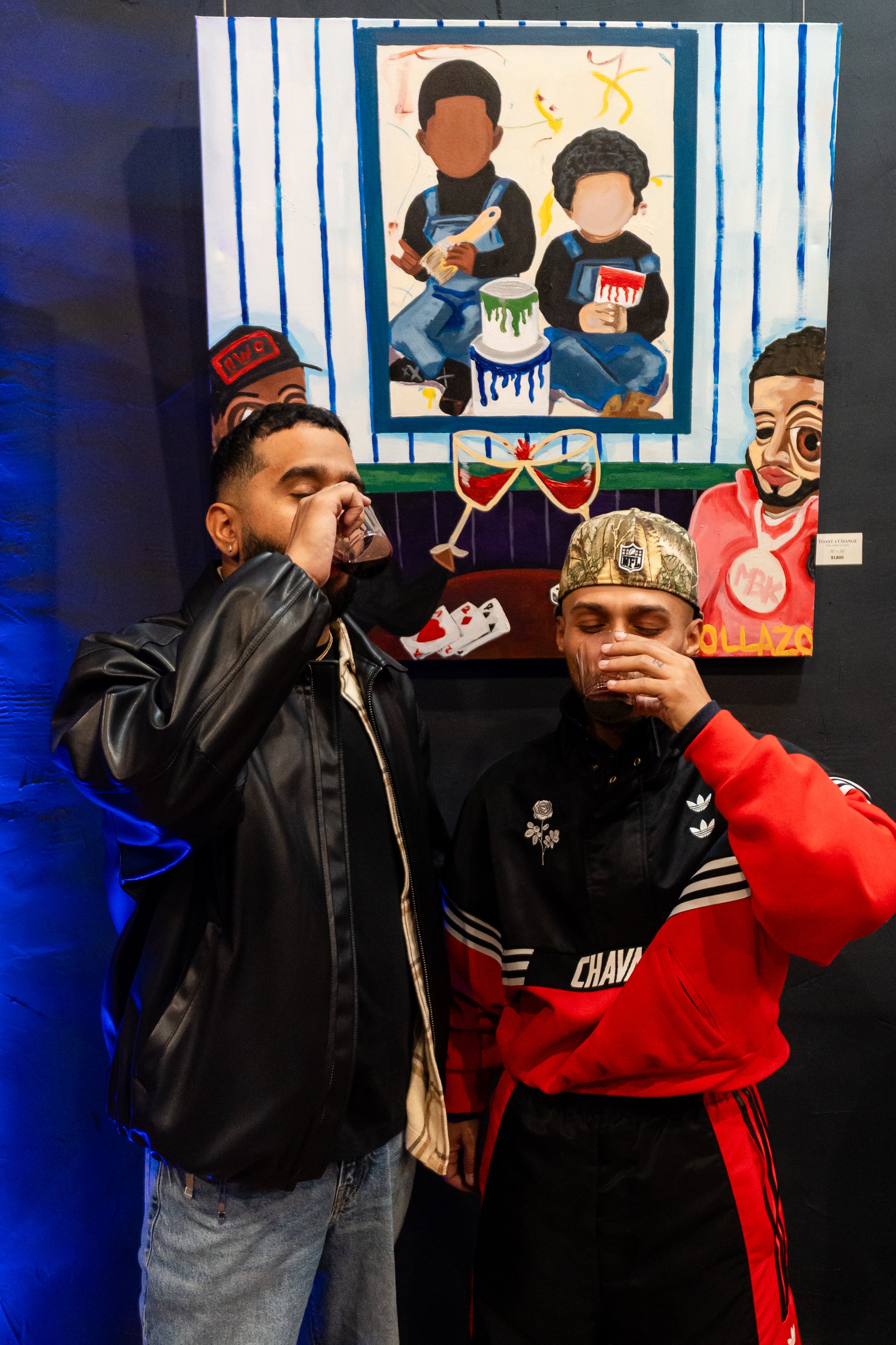 Two men are standing in front of an art piece, each holding a glass and drinking. The art painting behind them depicts two children, one holding a book and the other a cupcake, with a colorful, playful background.