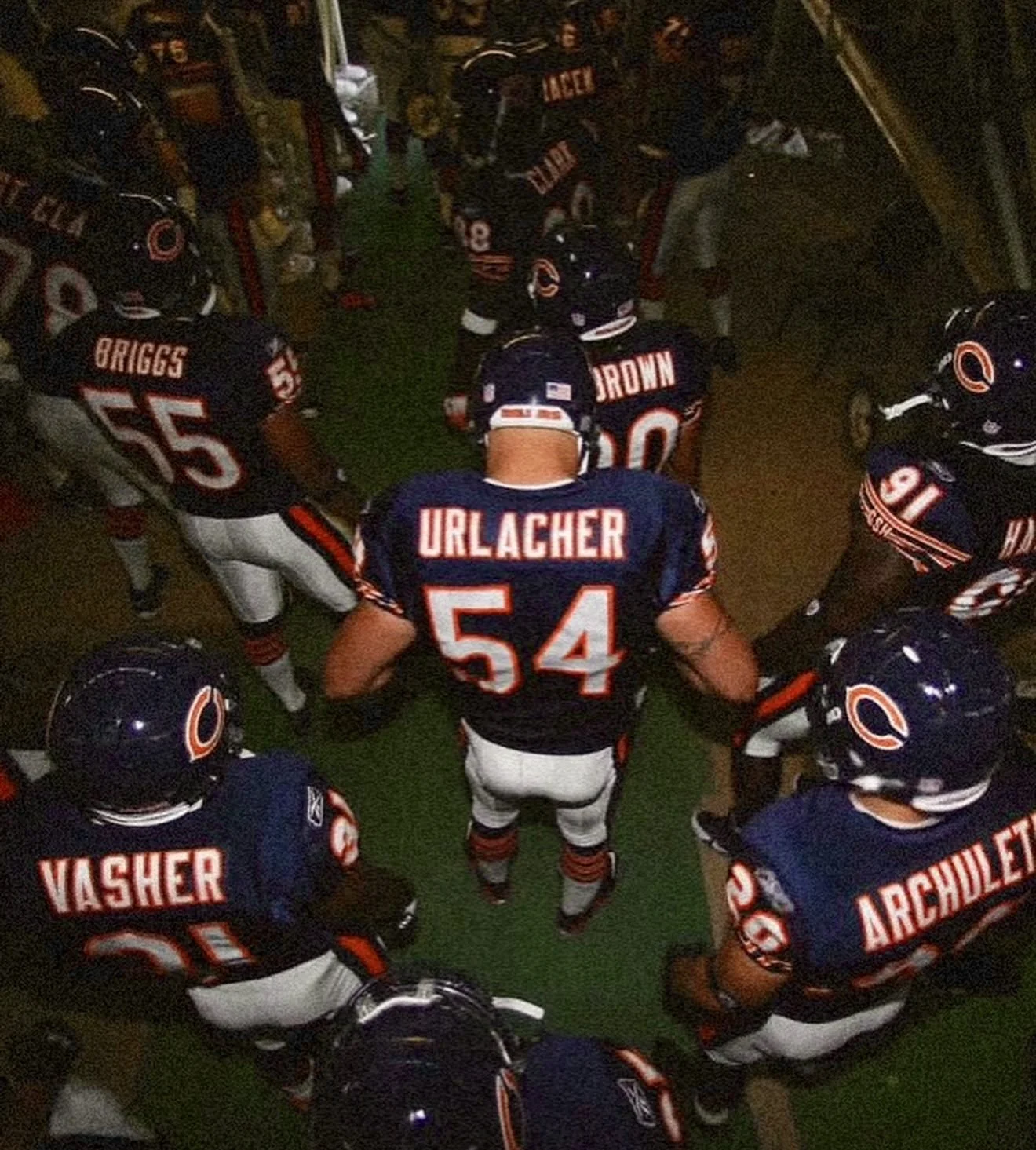A group of professional American football players from the Chicago Bears team is gathered in a circle on a green indoor field, preparing for the game. They are wearing black helmets and navy blue jerseys with white and orange accents, with their name