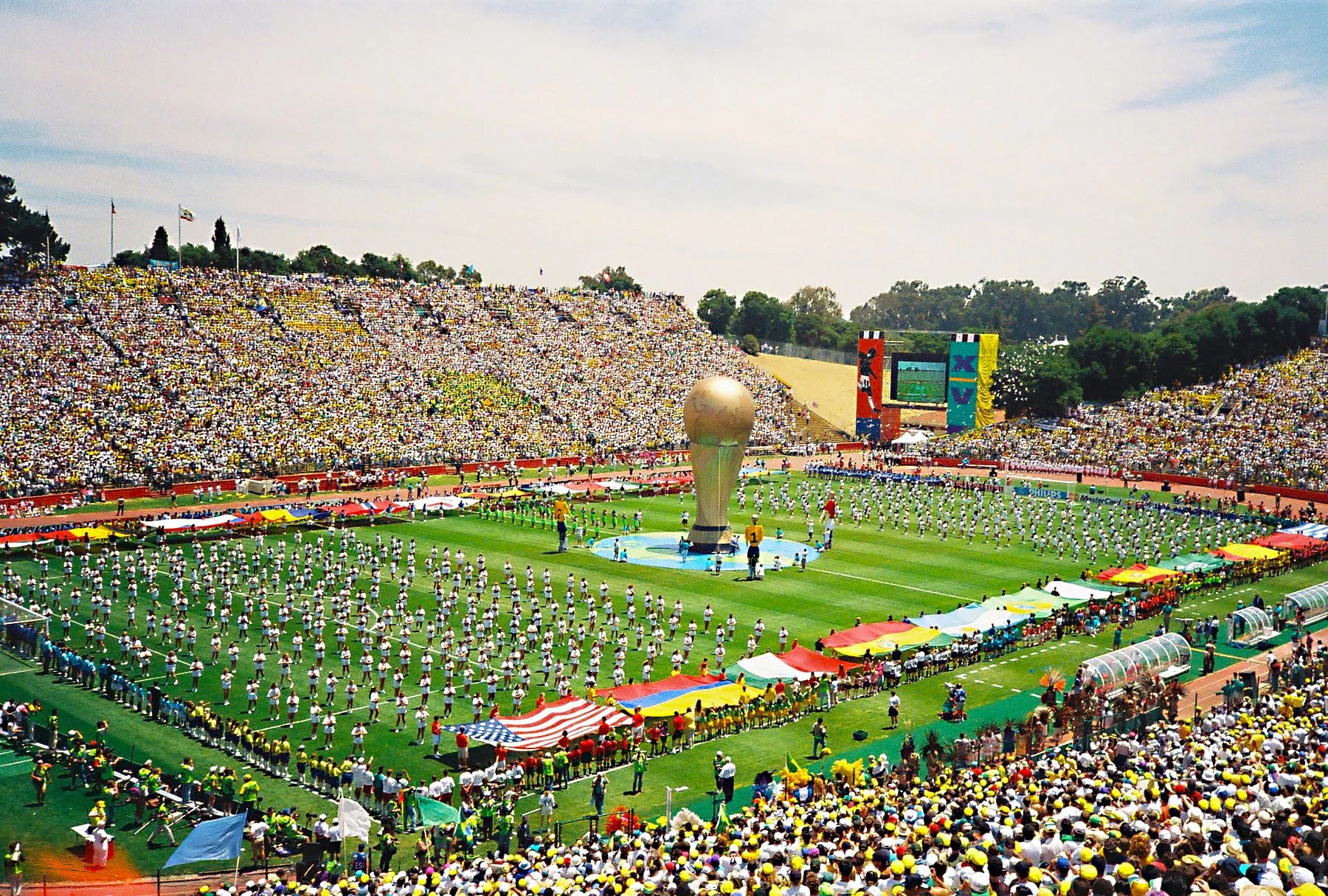 Crowd gathered at a stadium for a large international soccer event with a giant trophy as the centerpiece, surrounded by performers and flags.