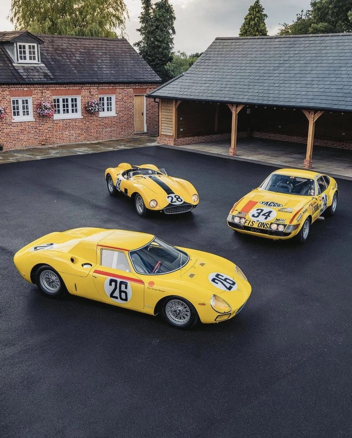 Three yellow vintage race cars parked on a black asphalt driveway with a brick house and a garage in the background.