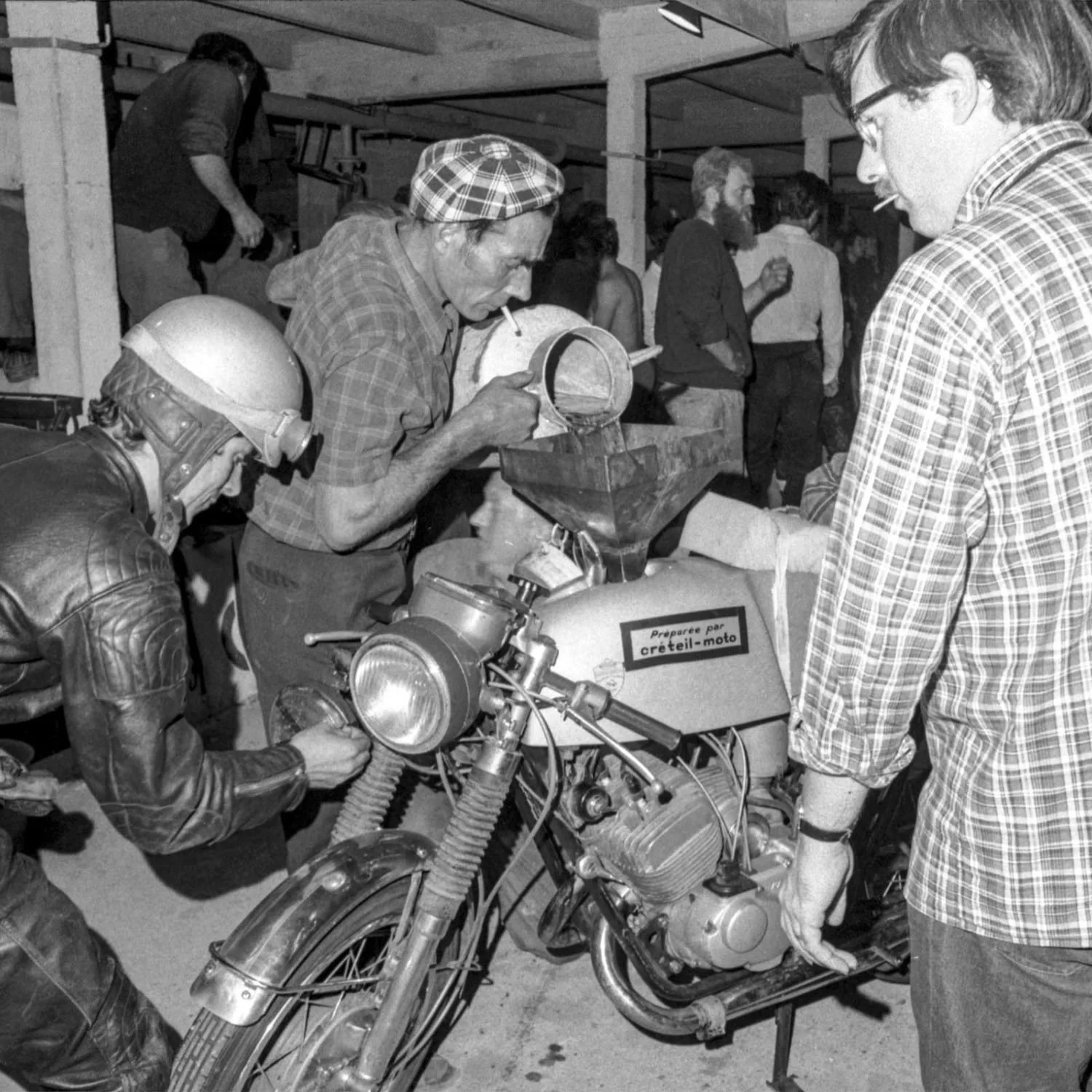 A black-and-white photo of three men working on a motorcycle inside a workshop, with several people standing and talking in the background.