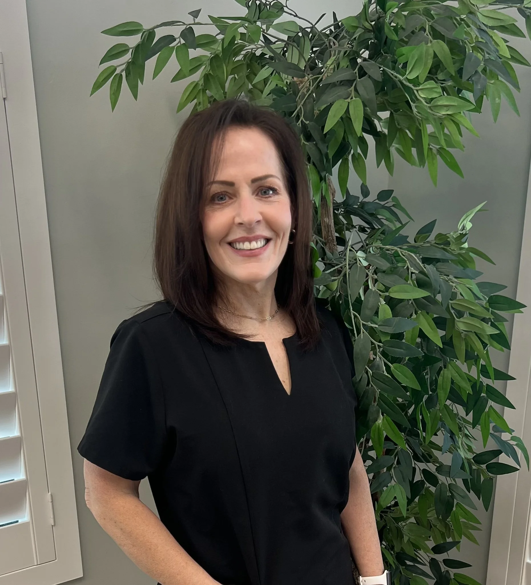 Smiling woman with dark hair wearing a black top and white watch, standing indoors next to a large green leafy plant and gray wall.