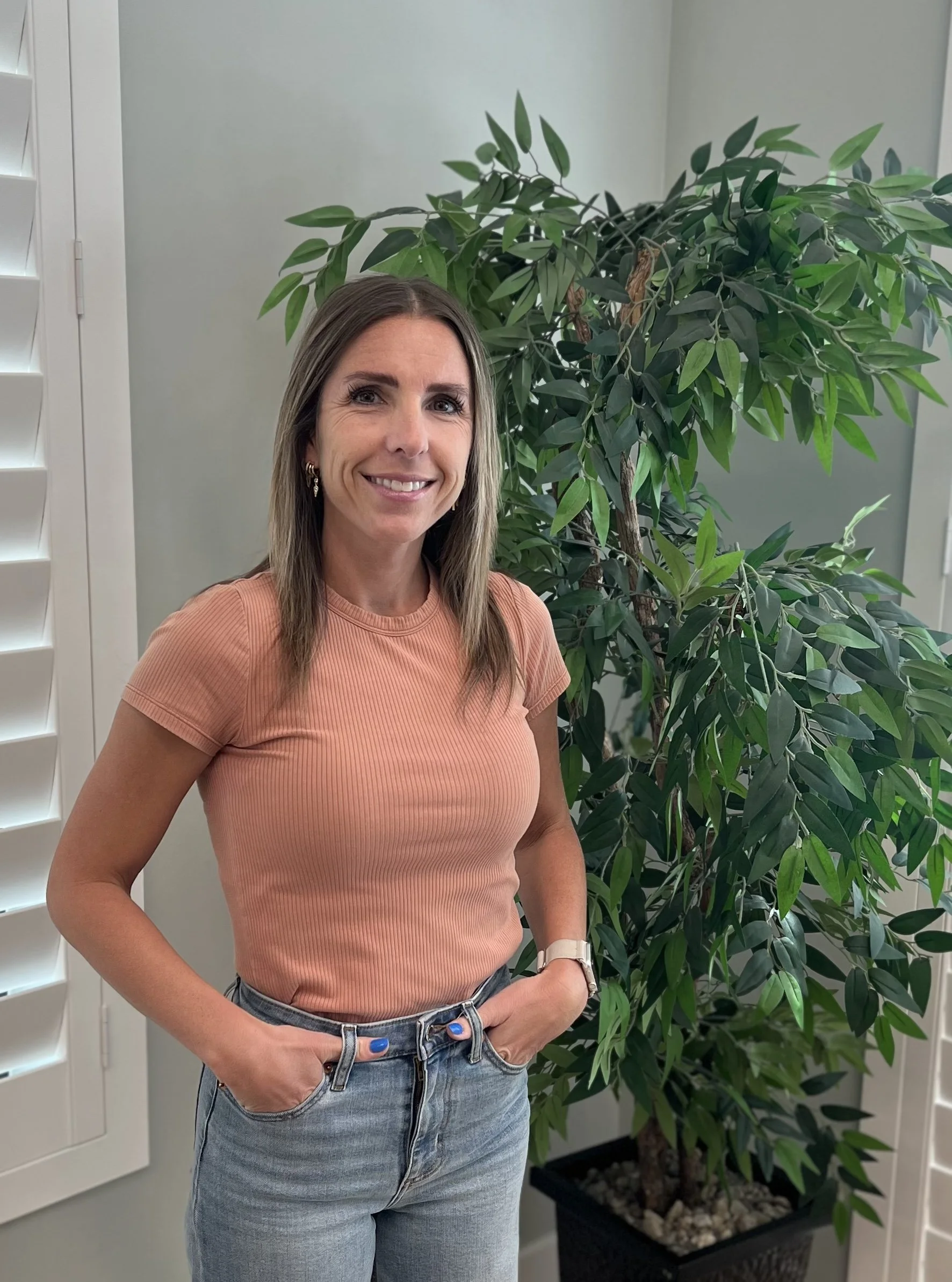 A woman standing next to a large leafy green plant indoors, wearing a peach-colored ribbed t-shirt, blue jeans, and a watch, with her right hand in her pocket and smiling at the camera.