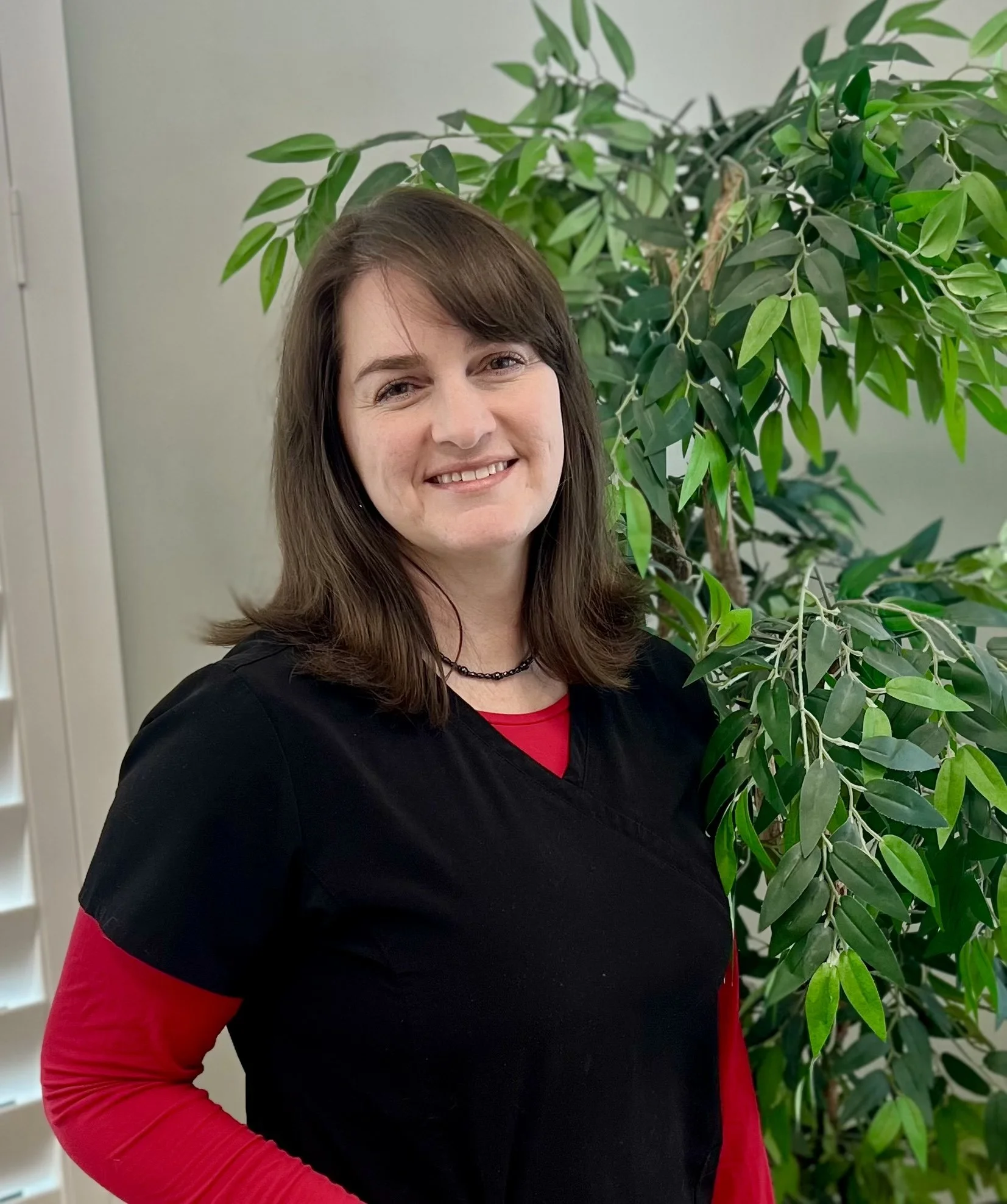 A woman with brown hair and a friendly smile, wearing a black top with red sleeves, standing in front of a large green leafy plant.