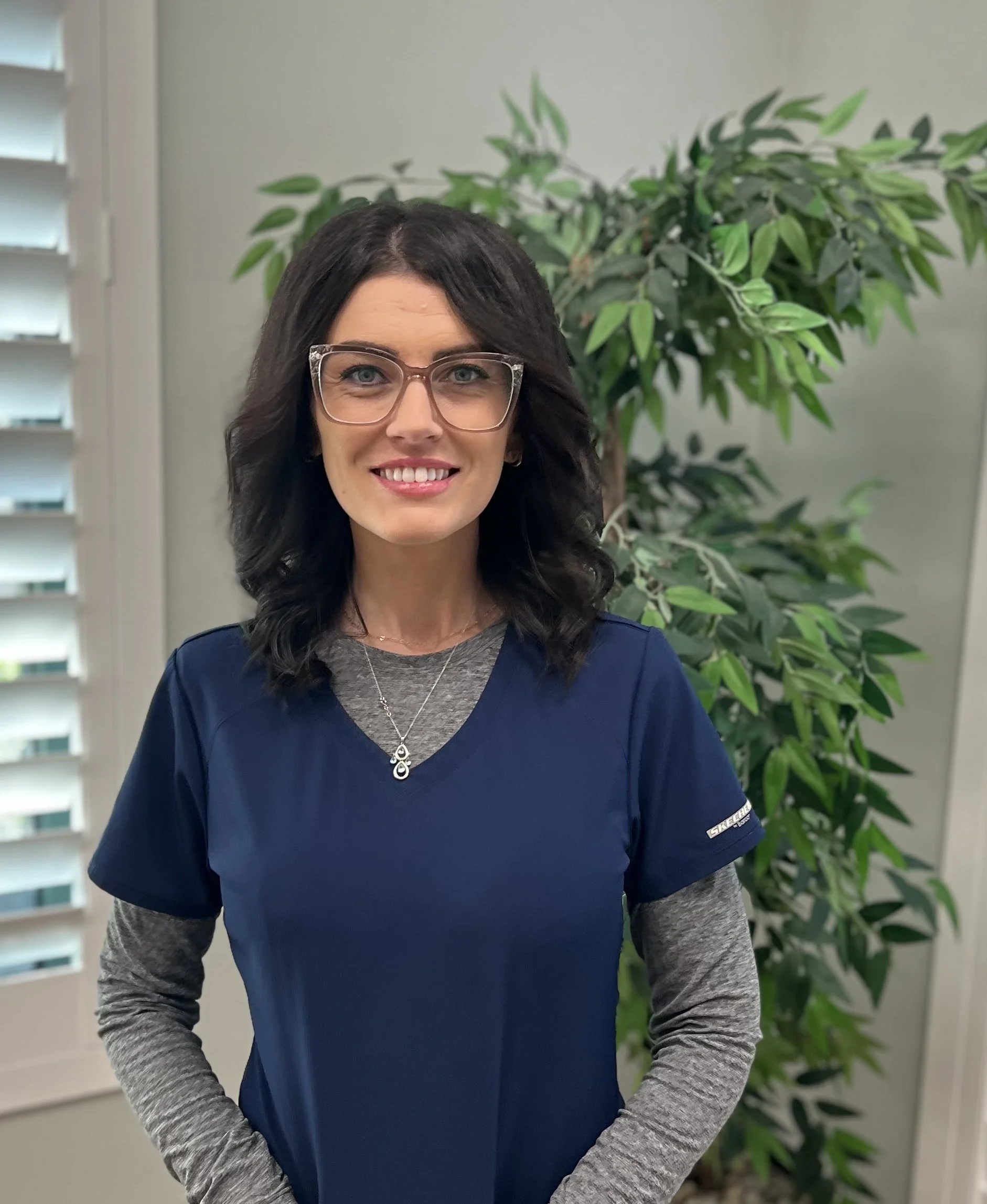 A woman with long dark hair, wearing glasses, a blue scrubs top over a gray long sleeve shirt, standing indoors with a green leafy plant and window blinds in the background.
