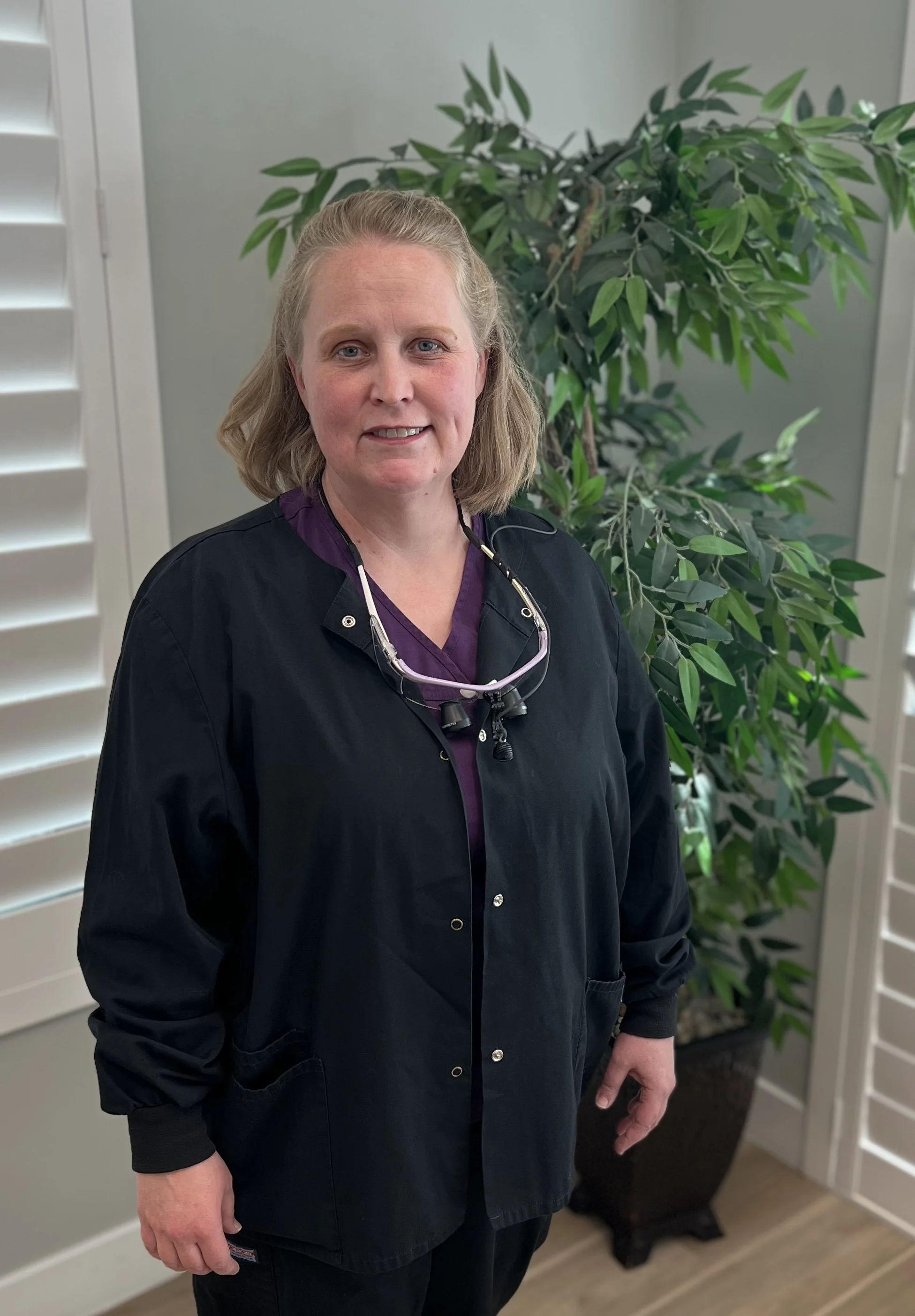 A woman with light brown hair wearing black medical scrubs and a stethoscope around her neck standing indoors near a large green leafy plant, with white shutters on a window behind her.