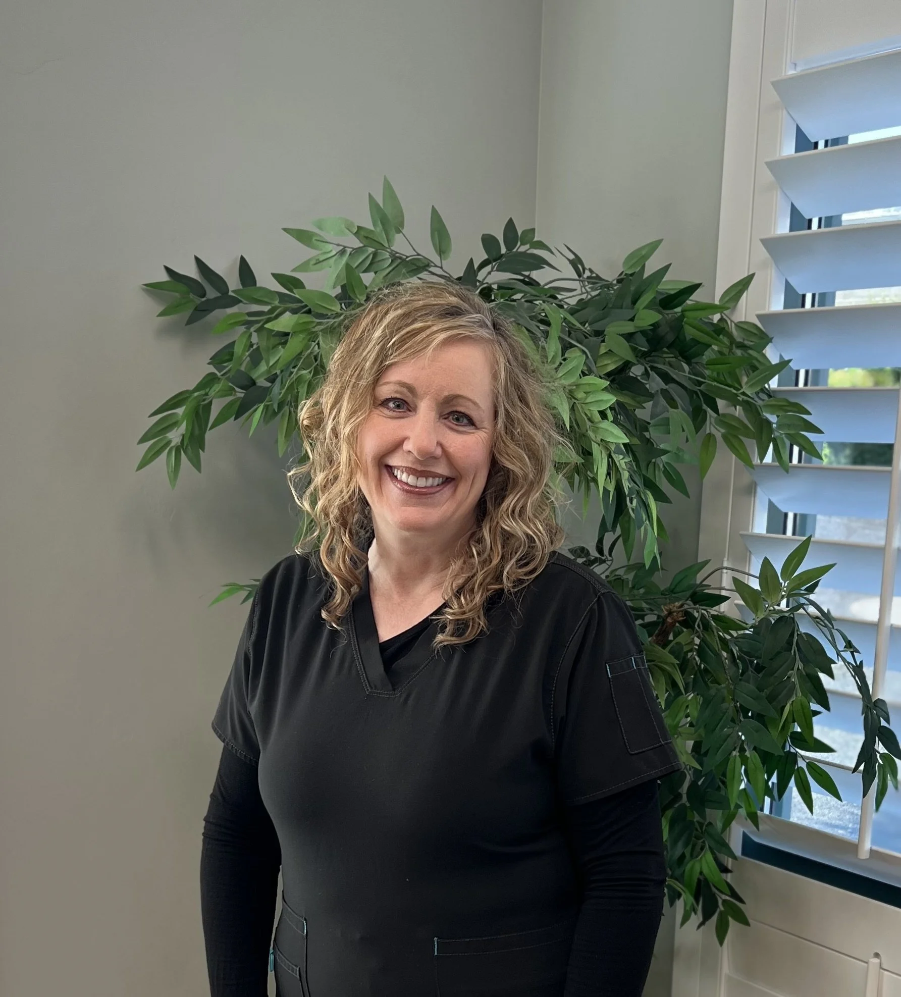 Smiling woman with curly blonde hair standing indoors near a large leafy plant, window with horizontal blinds, in a well-lit room.