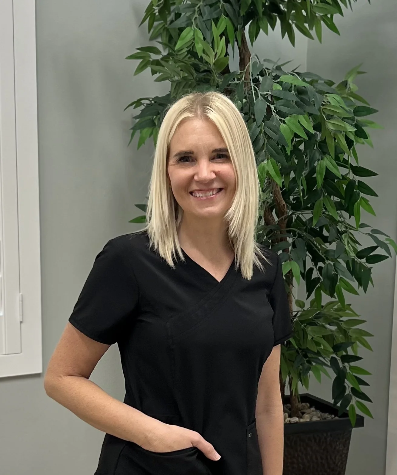 A woman with blonde hair wearing a black medical scrub top standing in front of a large green indoor plant.