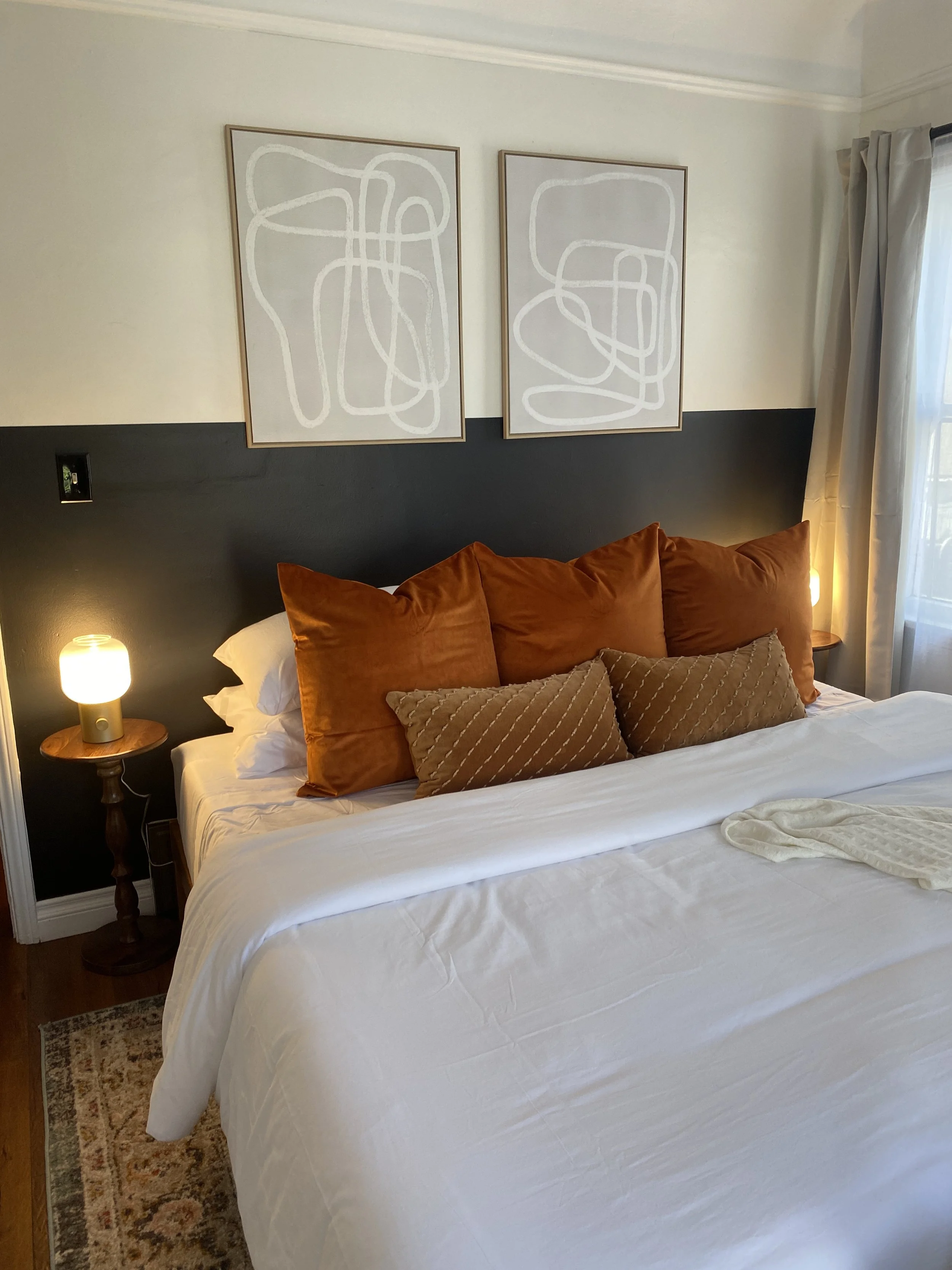 A cozy bedroom with a bed featuring large rust-colored pillows and smaller beige pillows, fronting a black accent wall with abstract artwork, side tables with lamps, and a window with white curtains letting in natural light.