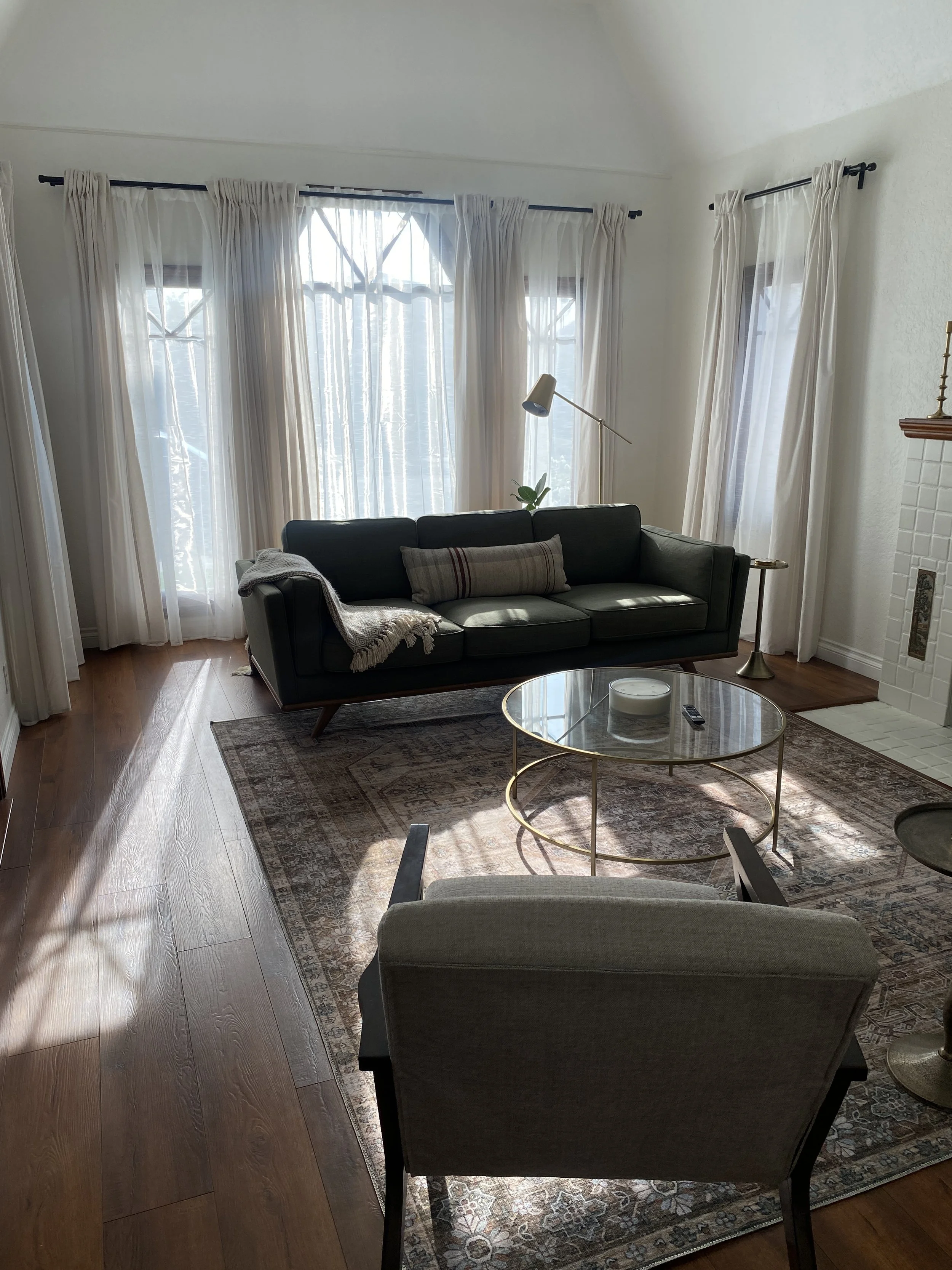 Living room with three large windows with white curtains, a dark green sofa with a beige pillow and throw blanket, a round glass coffee table with a candle and remote, a beige armchair, a patterned area rug, a floor lamp, and a fireplace with white t