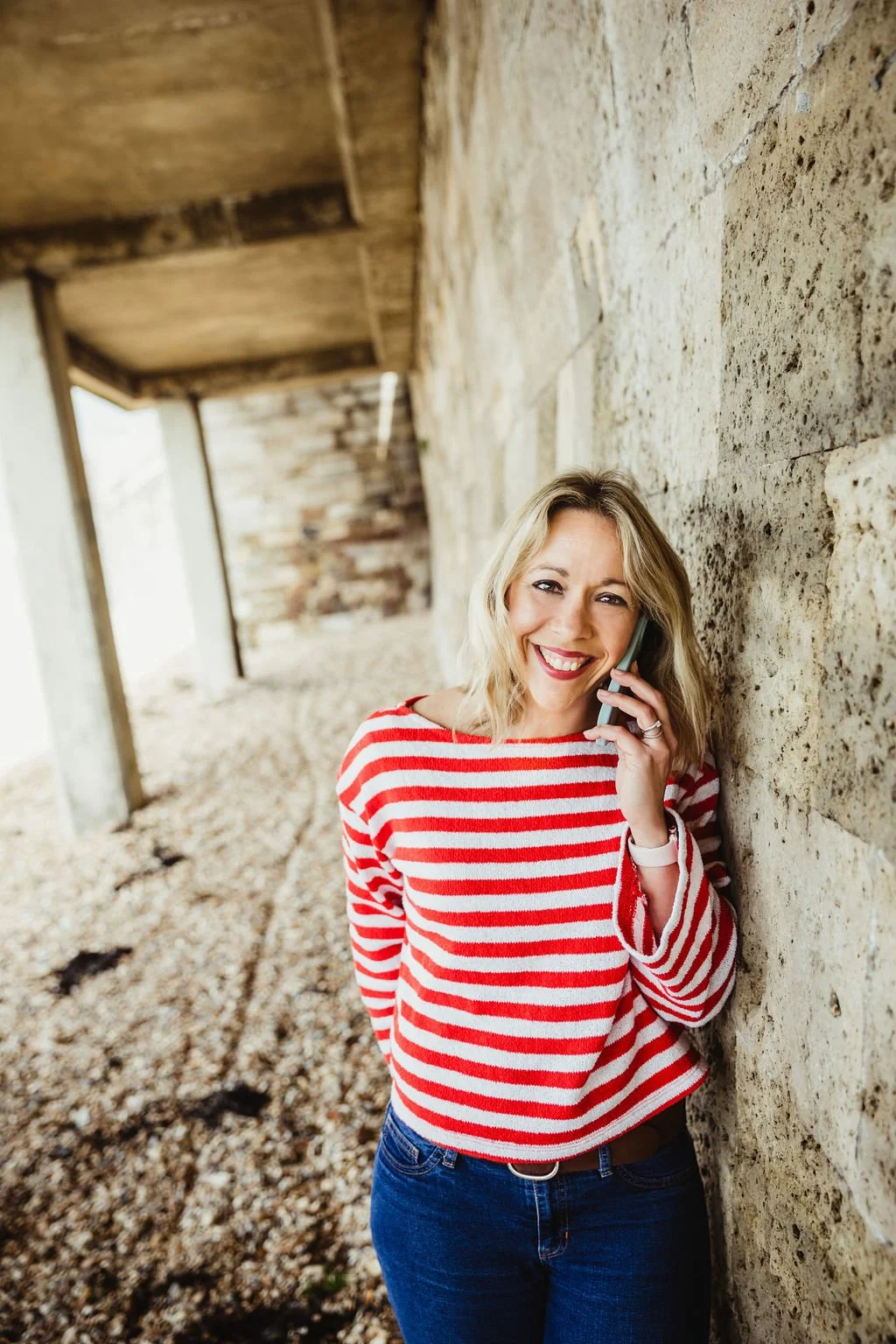 A woman with blonde hair, wearing a red and white striped sweater, talking on a cell phone and smiling, leaning against a stone wall under a wooden structure.