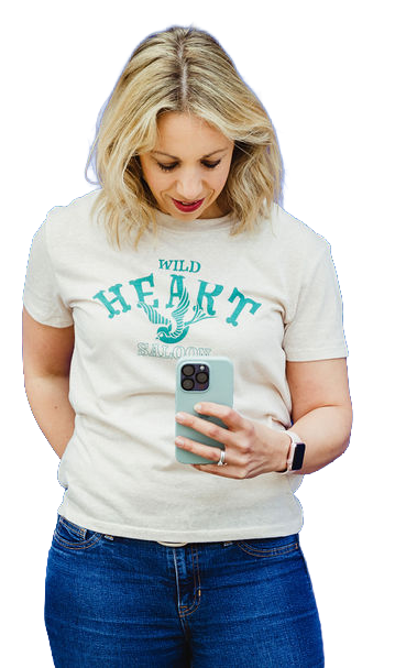 Woman taking a mirror selfie with her smartphone, wearing a white t-shirt that says 'WILD HEART' and blue jeans.