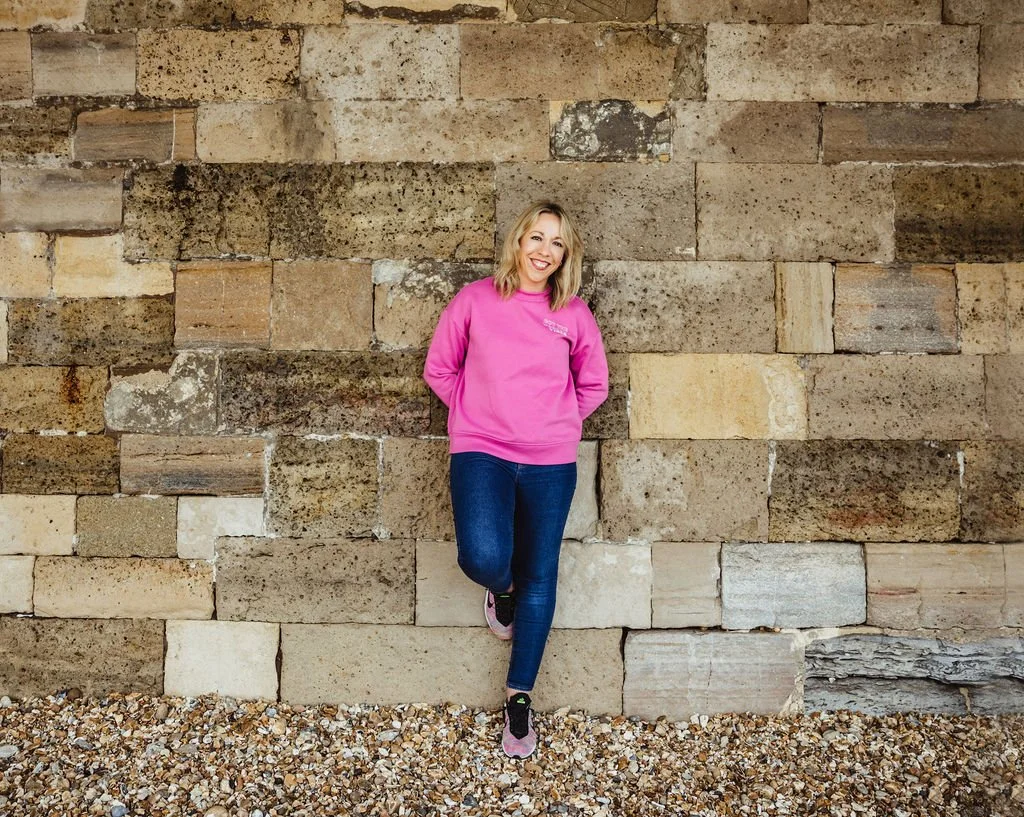 A woman with blonde hair wearing a pink sweatshirt and blue jeans standing against a stone brick wall, smiling and looking at the camera.