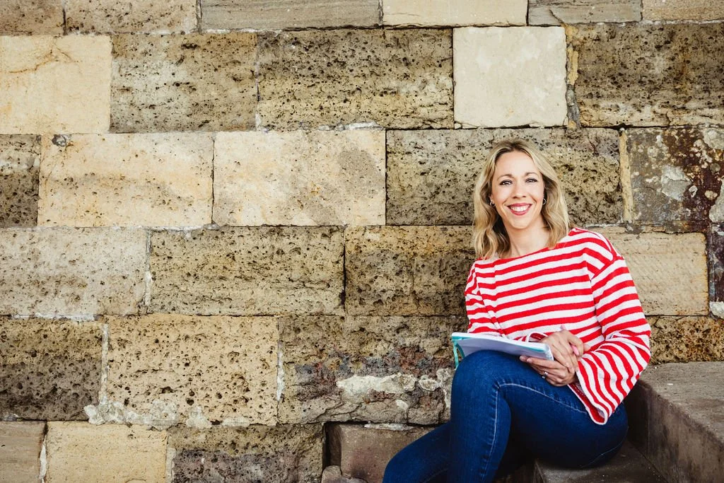 A woman with blonde hair, wearing a red and white striped shirt and blue jeans, sitting on steps against a stone wall, holding a notebook and smiling at the camera.