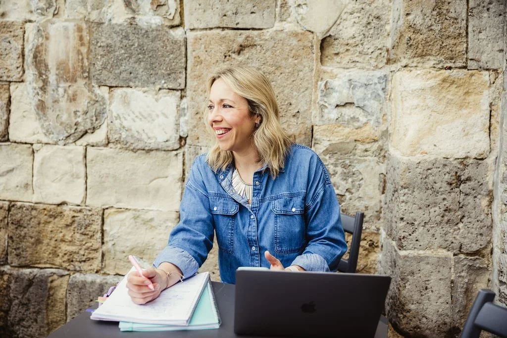 A woman with blonde hair, wearing a denim shirt, smiling and sitting at a table with a notebook and a laptop in front of her, against a stone wall background.
