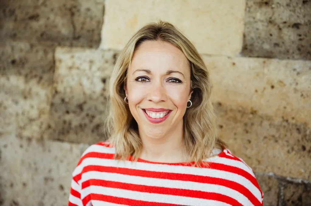 A woman with blonde hair smiling, wearing a red and white striped shirt, standing outdoors with a stone wall in the background.