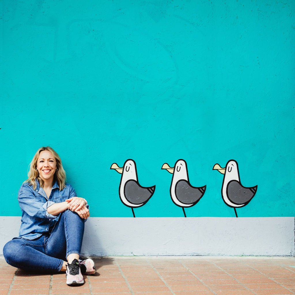 Woman sitting on the floor smiling next to a colorful wall with a mural of three cartoon seagulls.