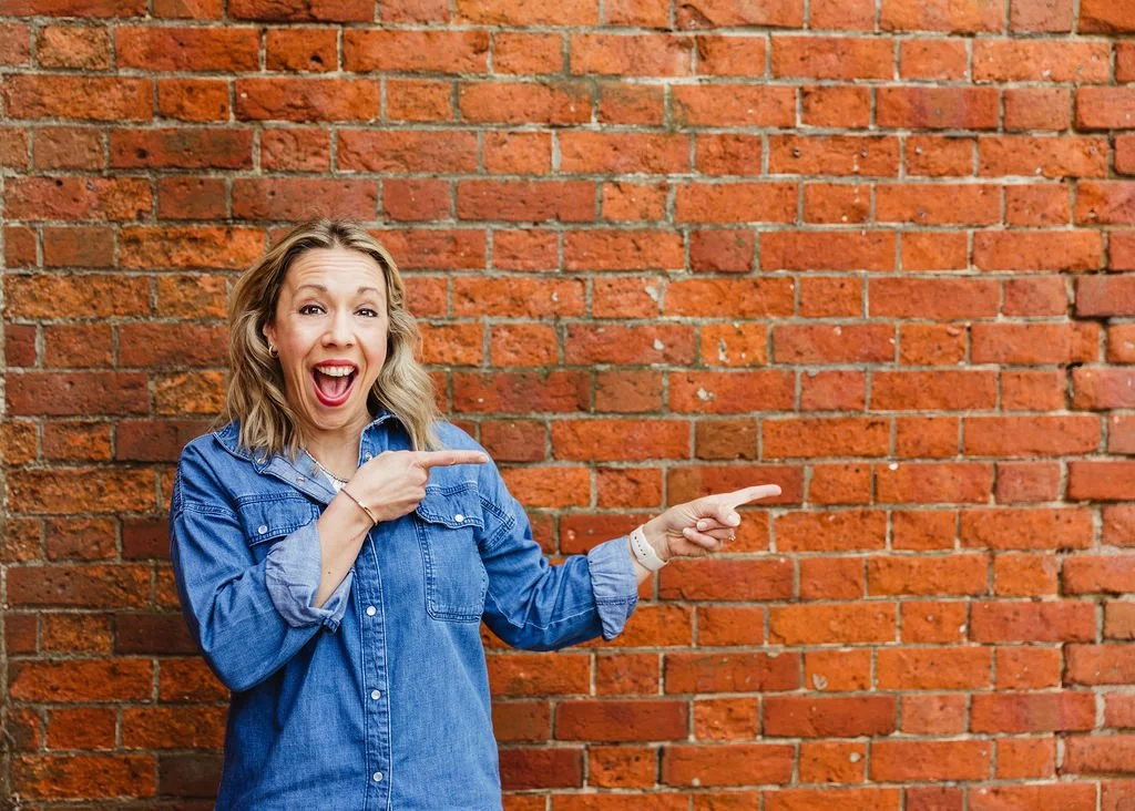 A woman with blonde hair smiling and pointing to the right while standing in front of a red brick wall.
