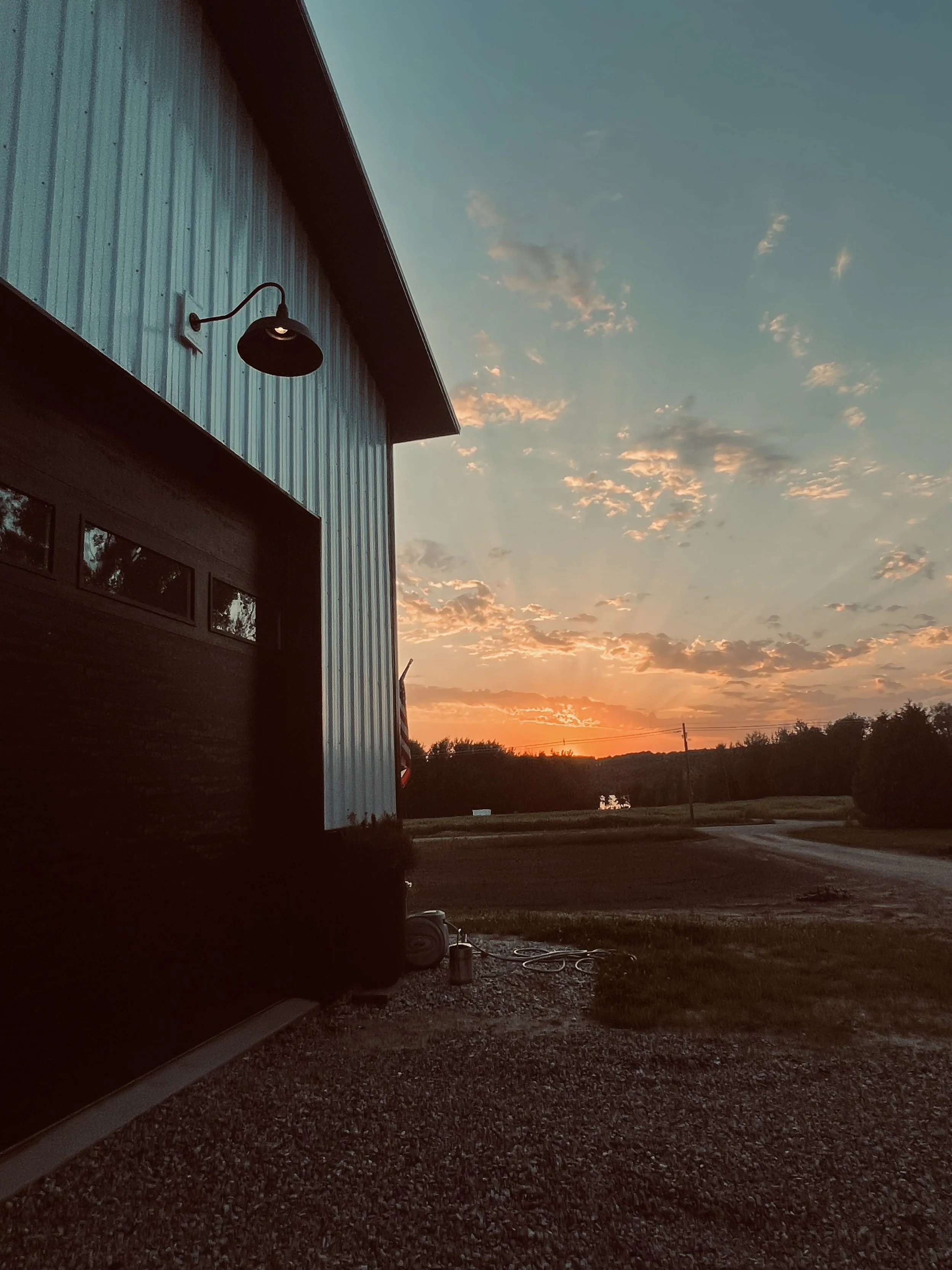 Barn exterior at sunset with light fixture, gravel driveway, and countryside background.