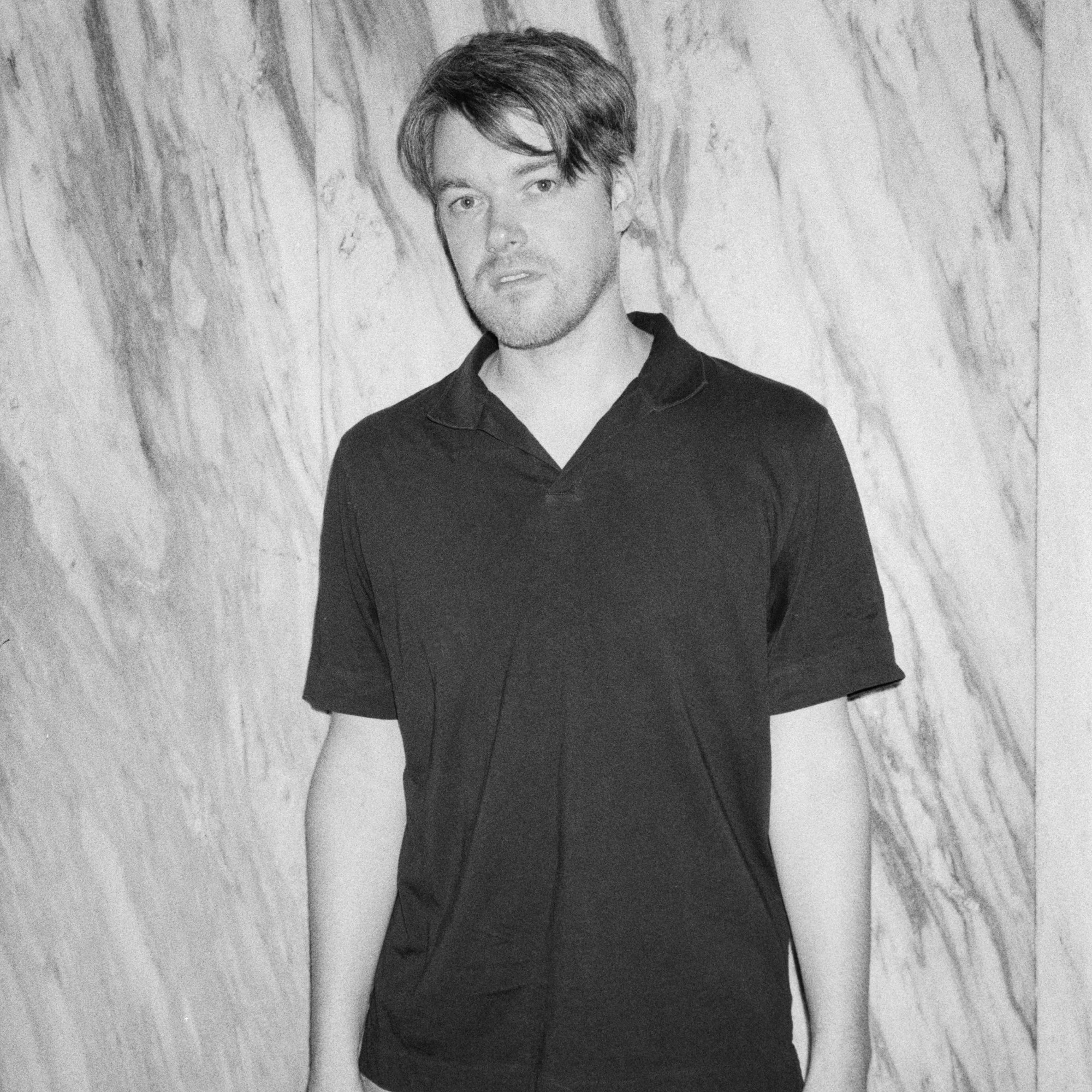 A young man with blonde hair wearing a black shirt and dark pants, standing against a light-colored background.