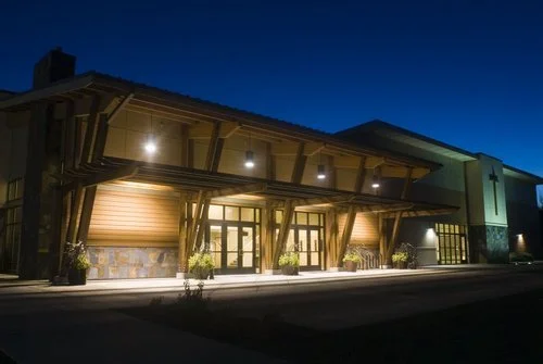 View of a modern building entrance at dusk with wooden structural elements, glass doors, and exterior lighting.