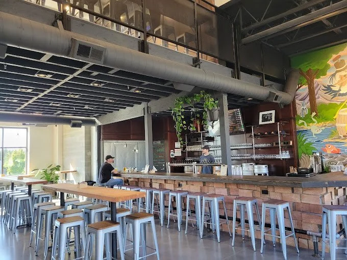 Modern cafe interior with a long bar counter, metal stools, a mural wall, and large windows providing natural light.