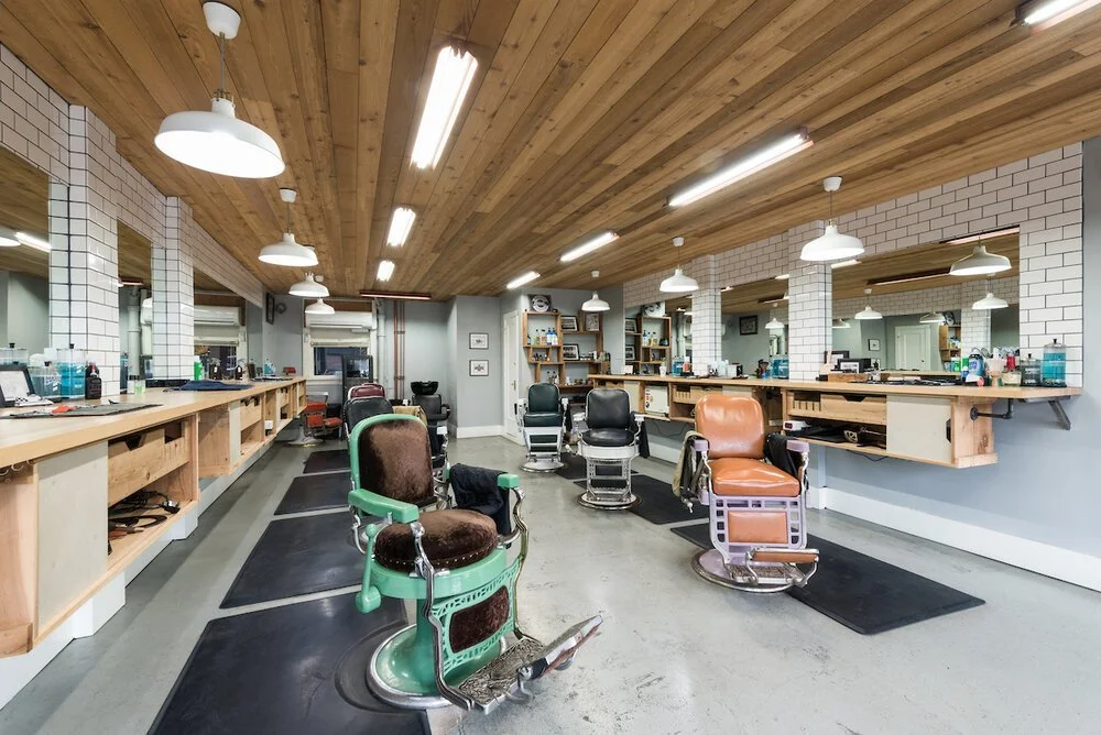 Barber shop with vintage barber chairs against a wall with mirrors and wooden shelves, illuminated by ceiling lights and pendant lamps.