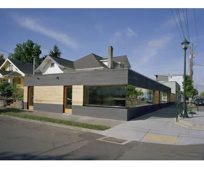 A modern, single-story building with large glass windows and a light-colored facade, situated on a corner lot in a suburban area with trees and utility poles nearby.