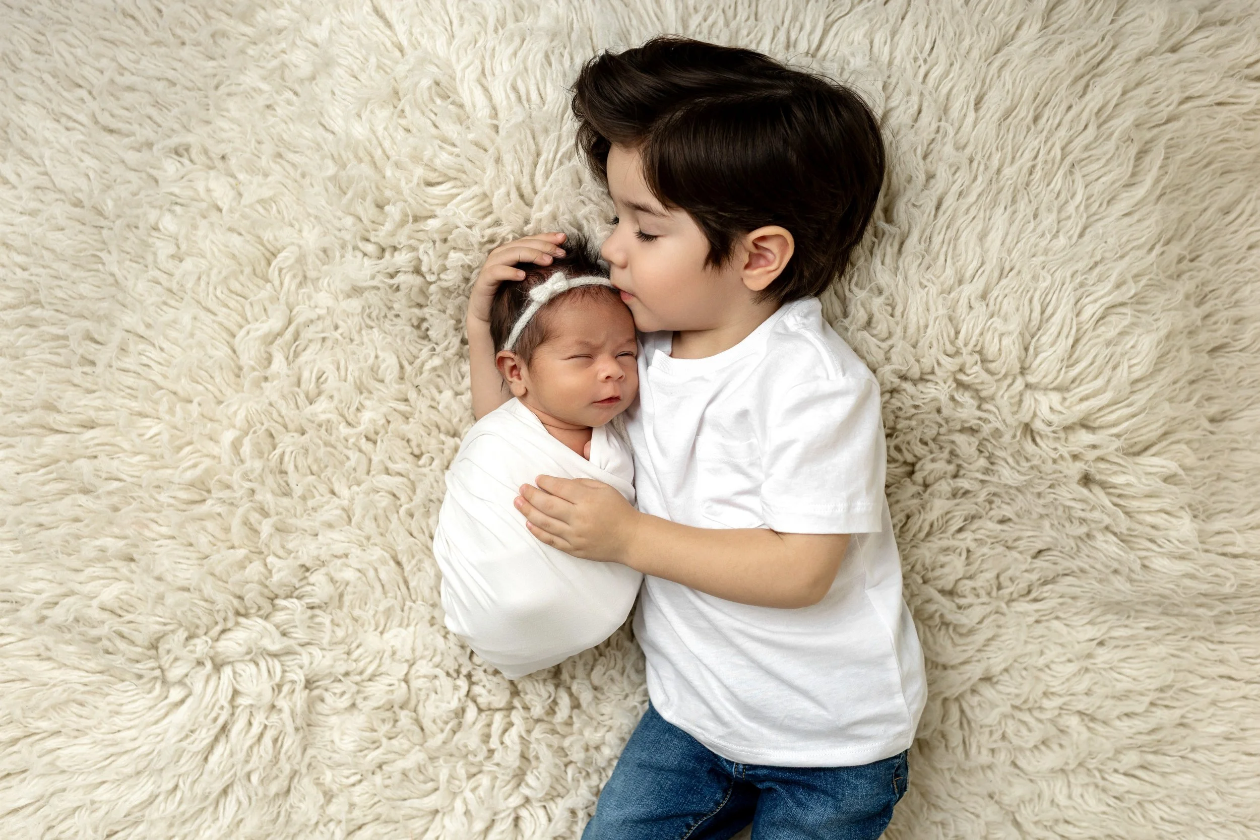 Older brother cuddling and kissing his newborn sister during a newborn photography session in Syracuse, Utah