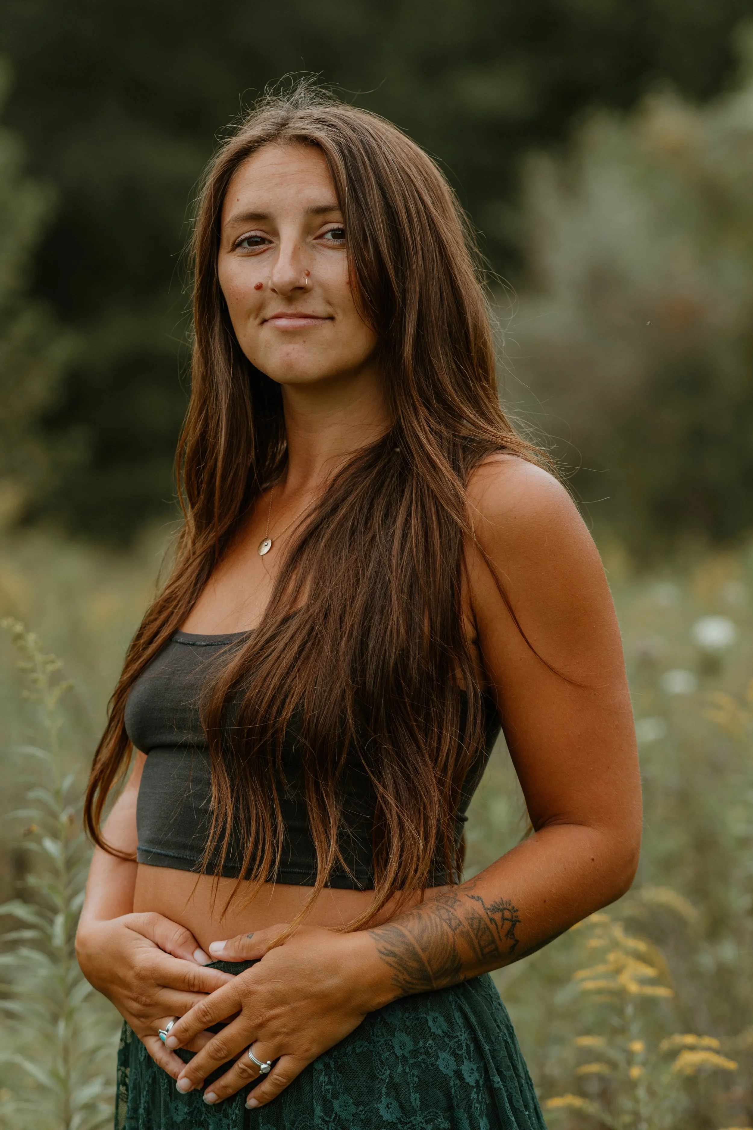 A young woman with long brown hair and tattoos on her arm, standing outdoors in a field of tall plants with a blurred natural background.