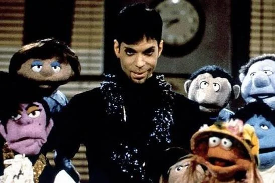 Prince performing Starfish and Coffee on Muppets Tonight, surrounded by Muppet characters in a classroom setting, 1997