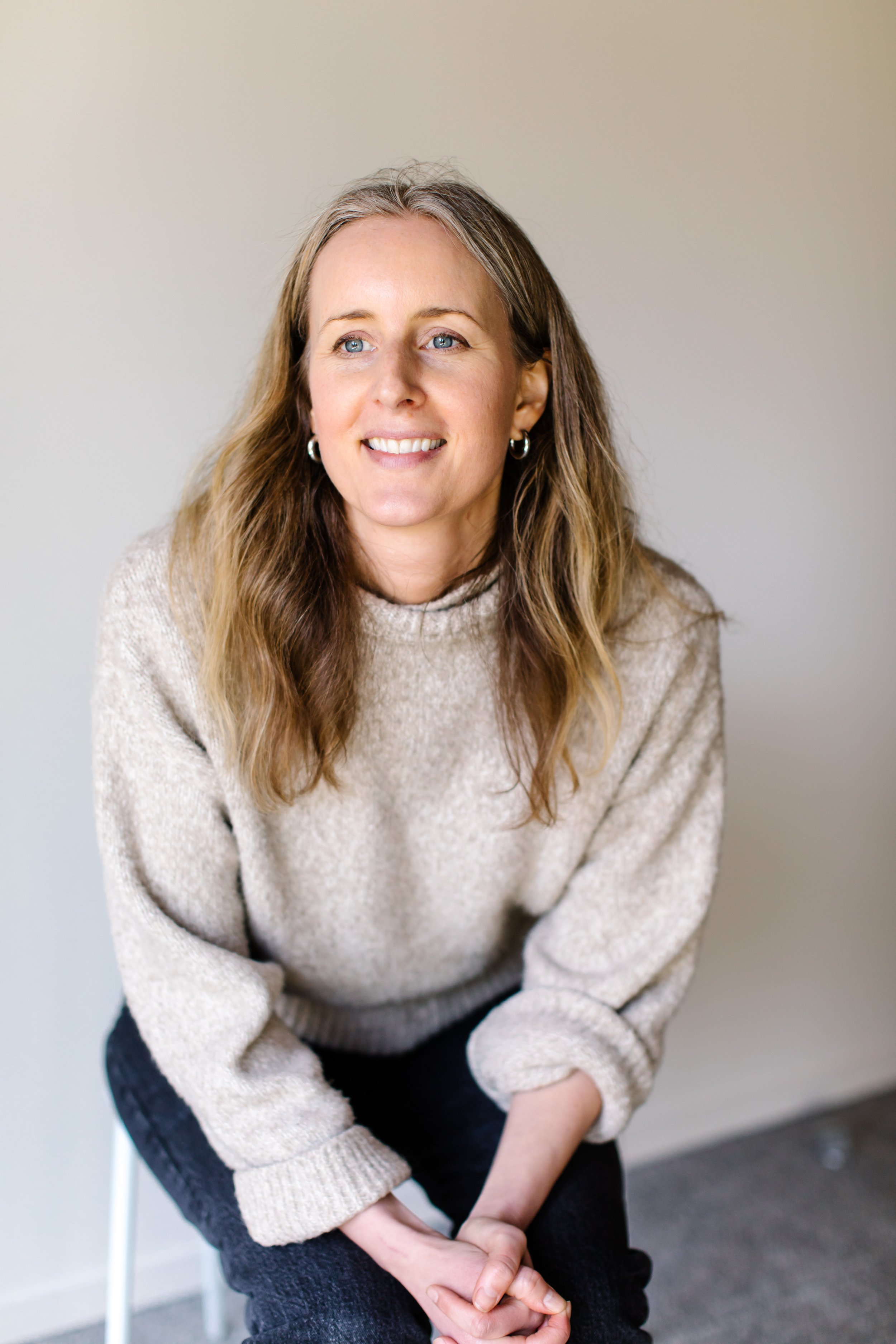 A person with long hair wearing a beige sweater and black pants, sitting and smiling.