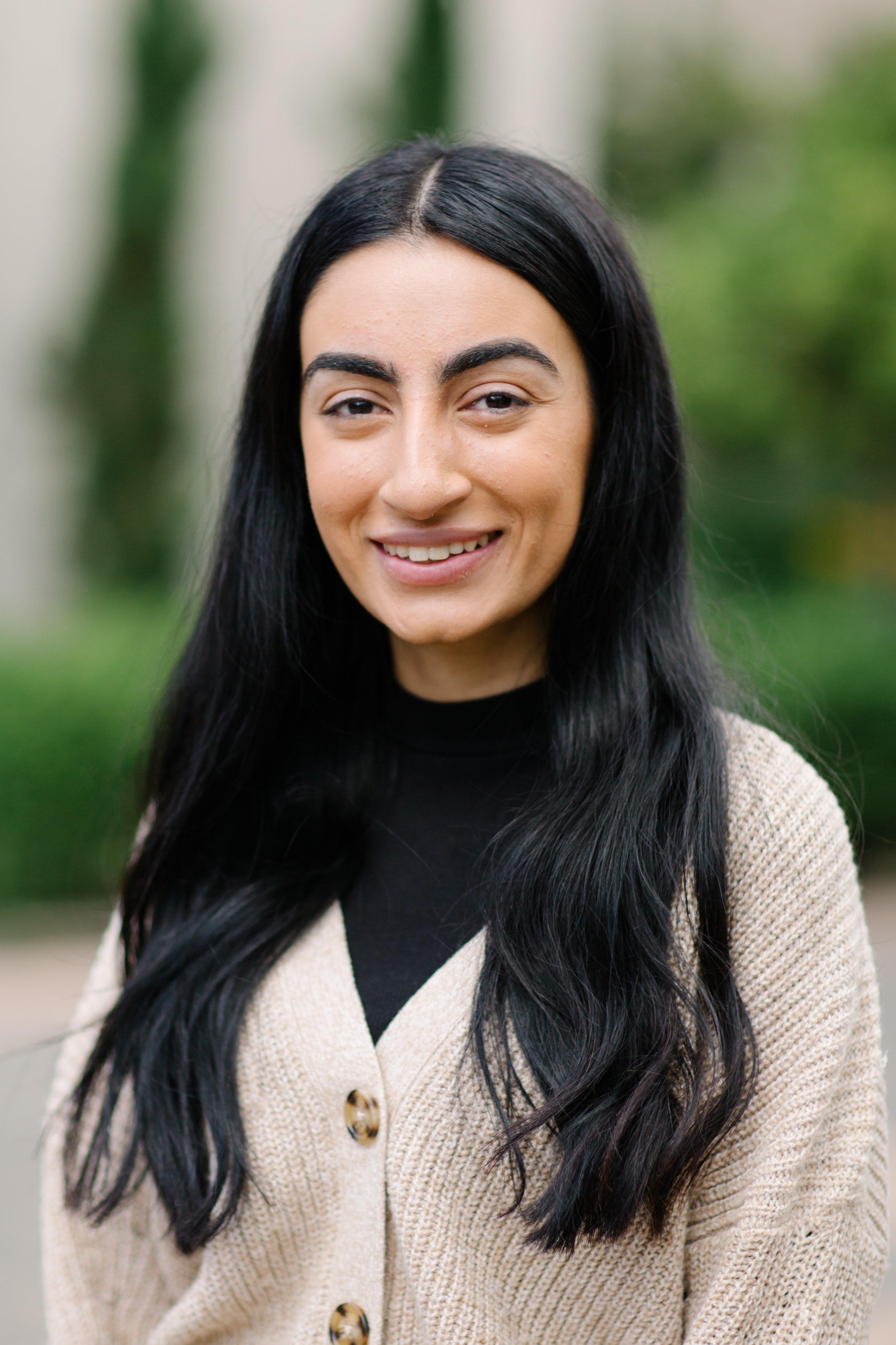 Smiling person with long black hair wearing a beige cardigan outdoors.