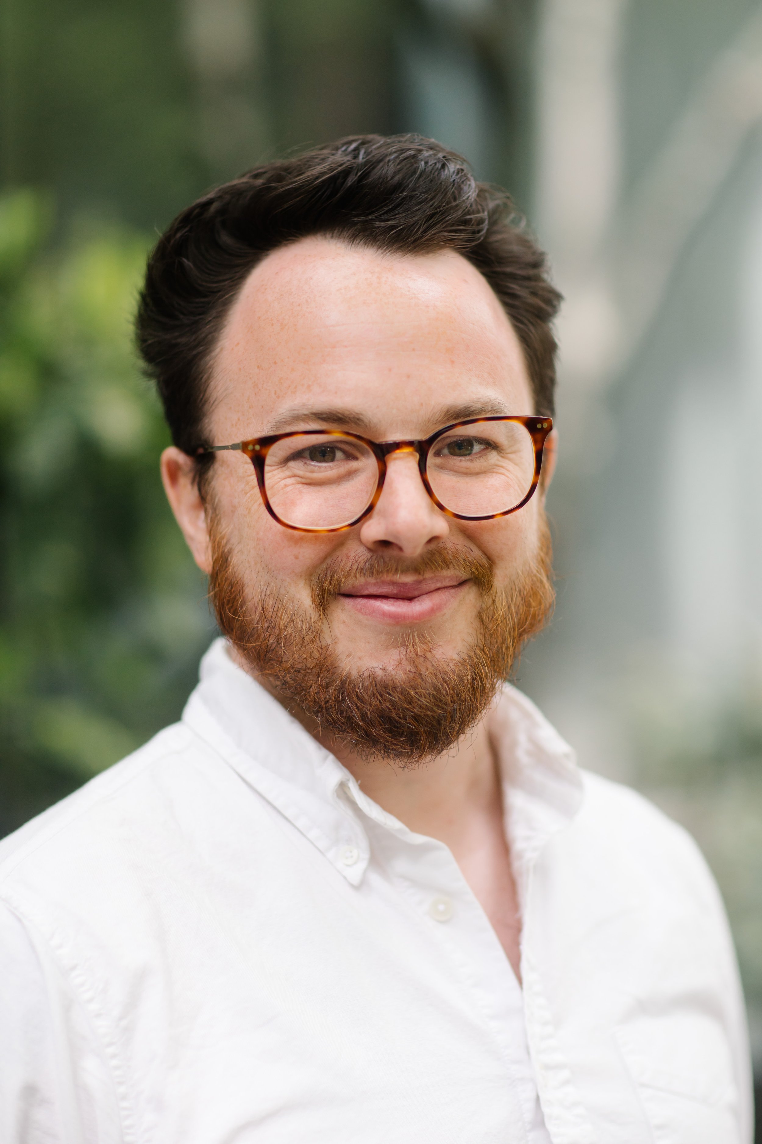 A man with glasses and a beard wearing a white shirt, smiling outdoors.