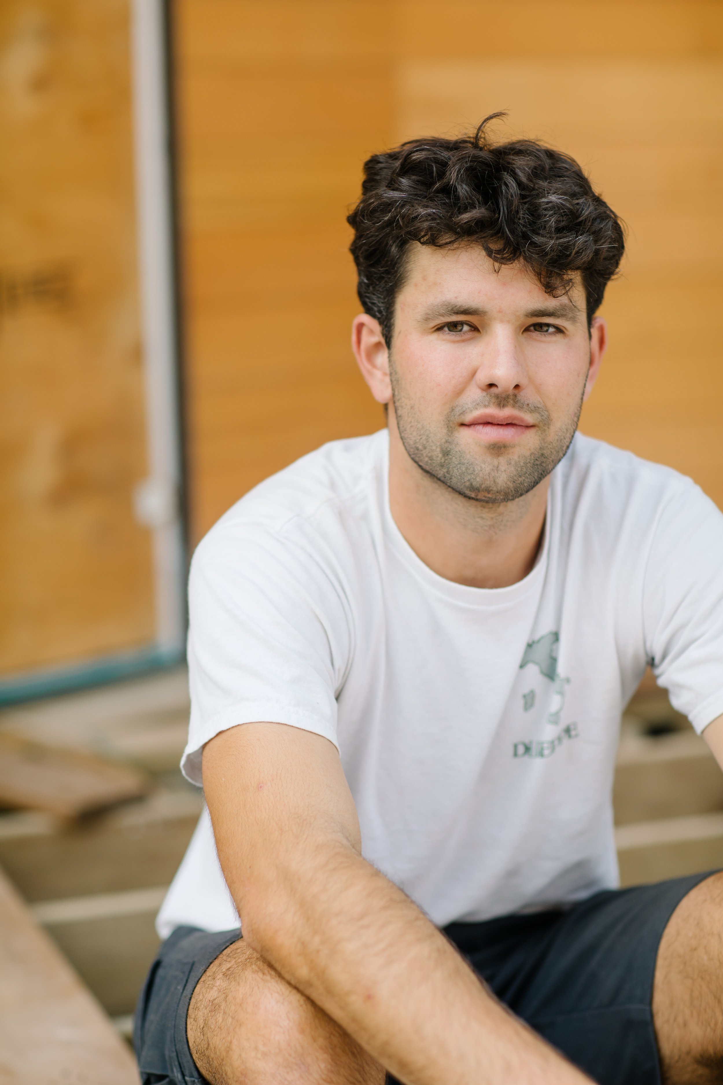 Man sitting outdoors wearing a white t-shirt and shorts, with a wooden background.