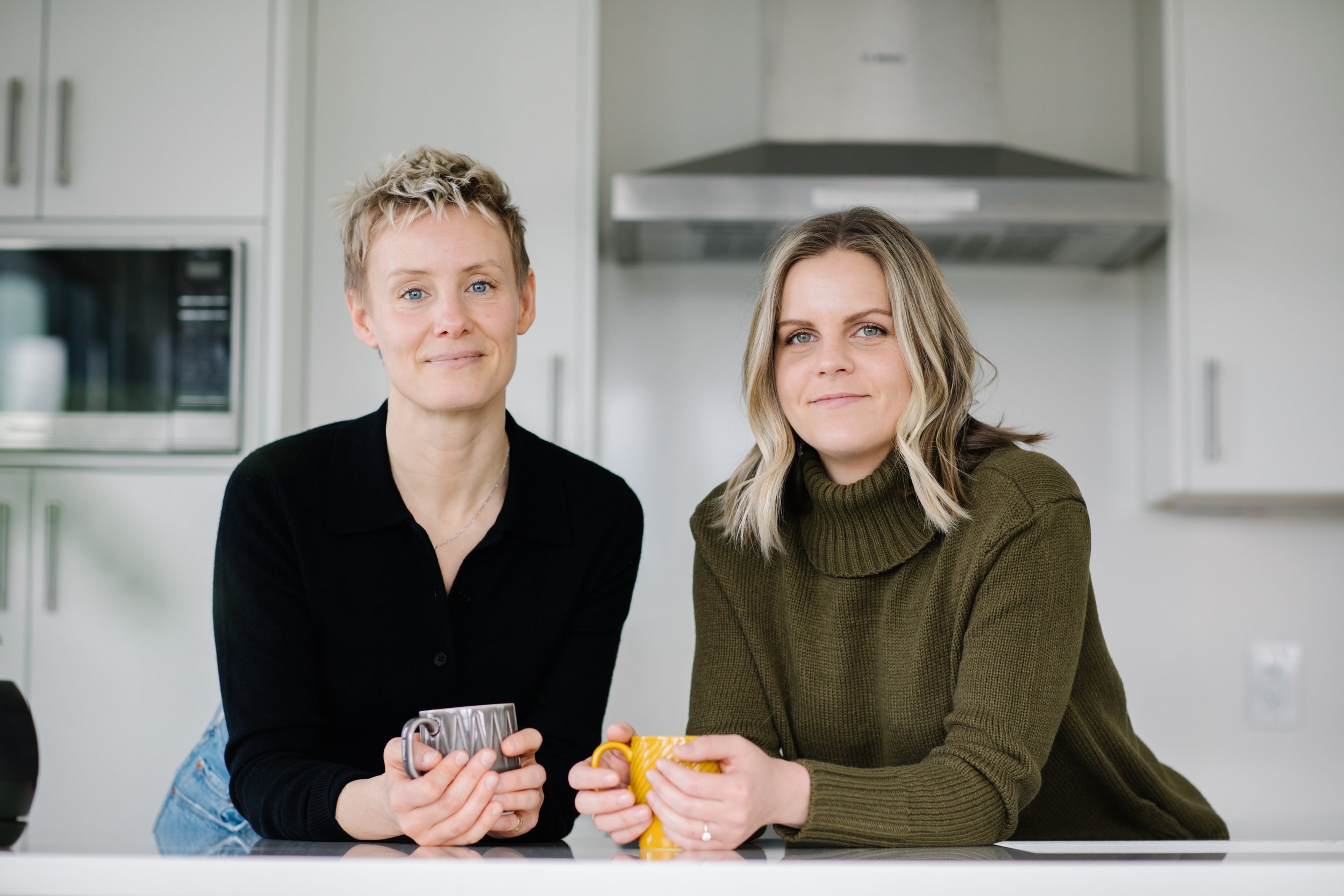 Two people in a kitchen holding mugs, one in a black top and the other in a green sweater, with a microwave and range hood in the background.