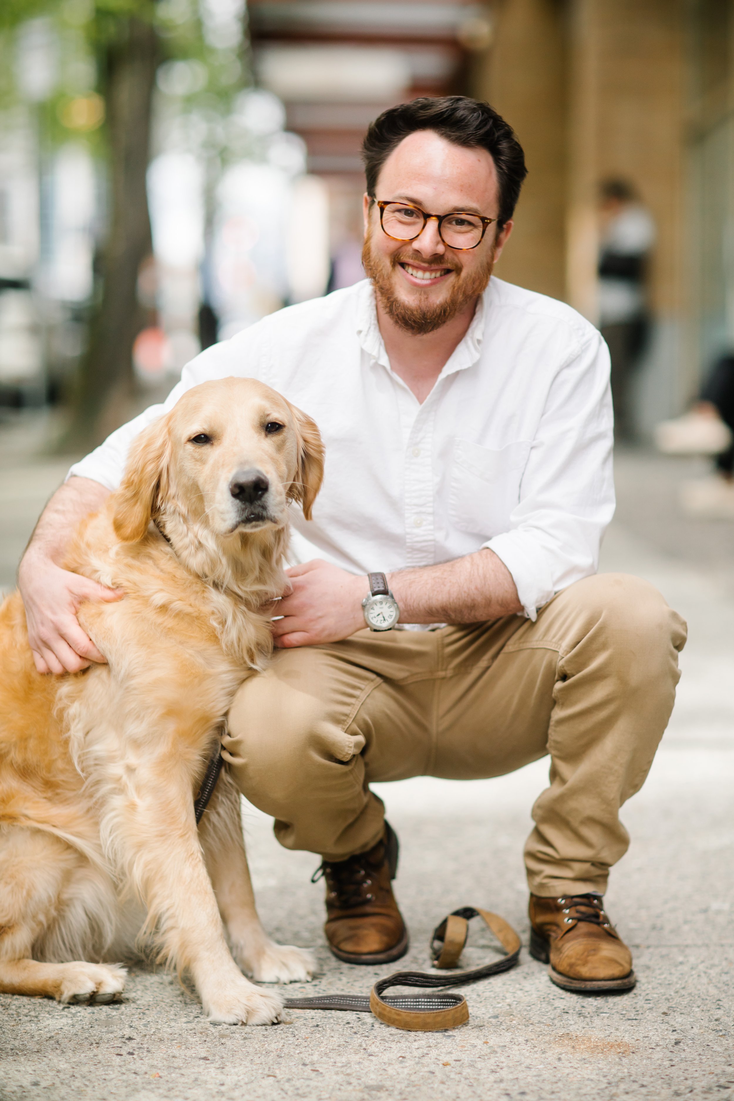 Man crouching on sidewalk with a Golden Retriever dog, both looking at the camera, with trees and buildings in the background.