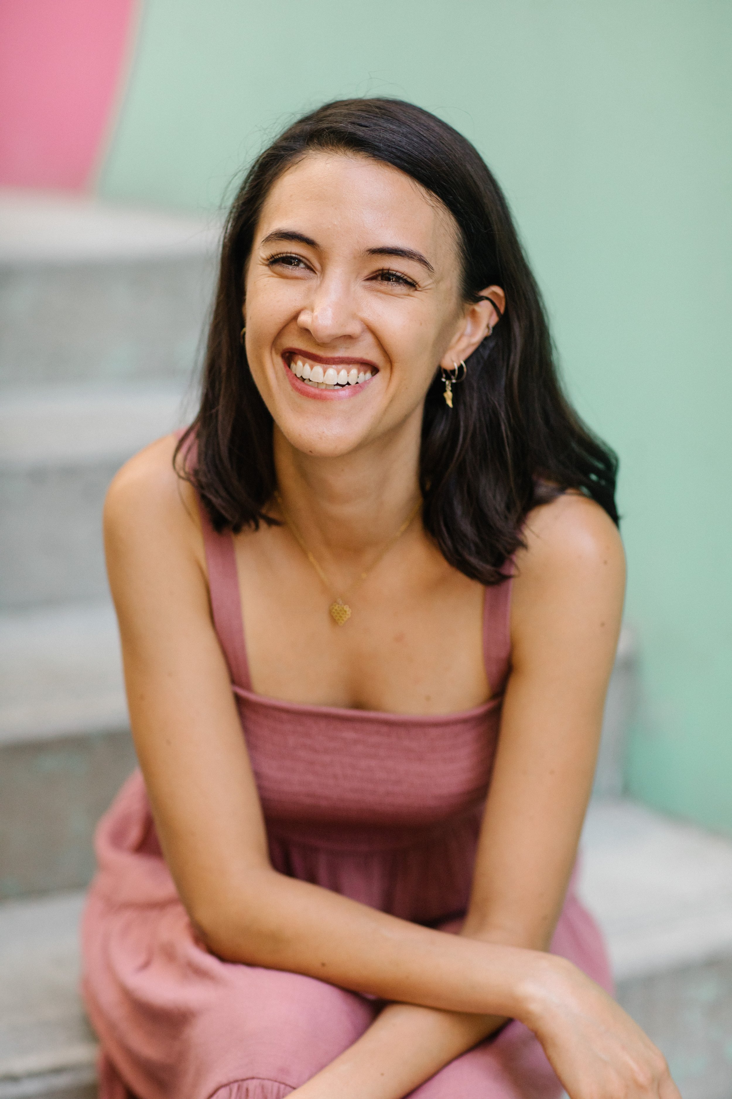 Smiling woman sitting on stairs with colorful background, wearing a pink dress and gold jewelry.