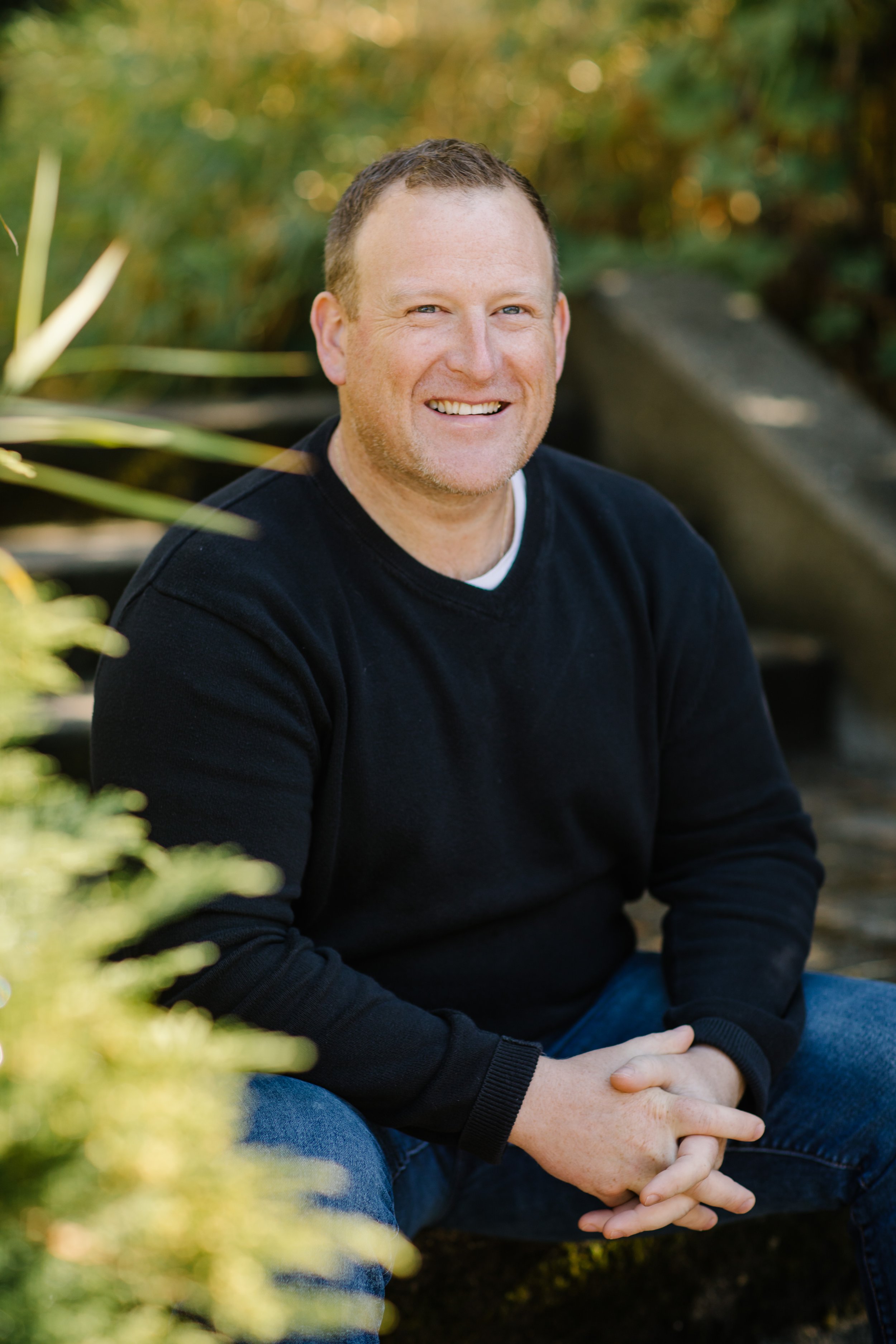 Smiling man in black sweater sitting outdoors in a garden.