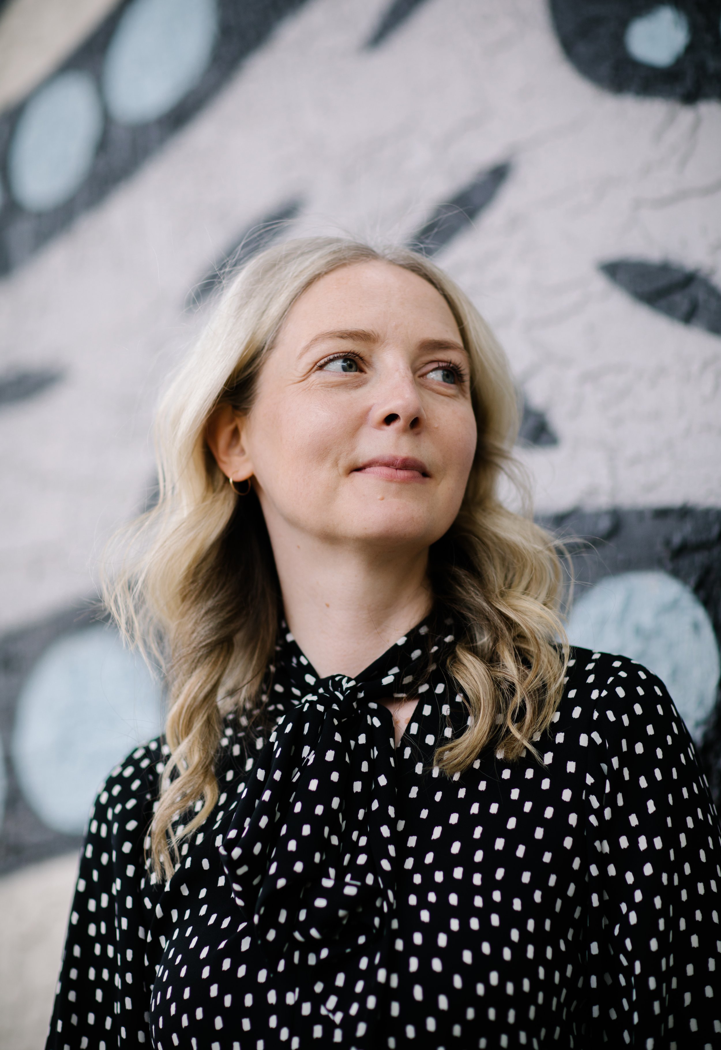 Person with blonde hair wearing a black and white polka dot blouse, standing in front of a mural.