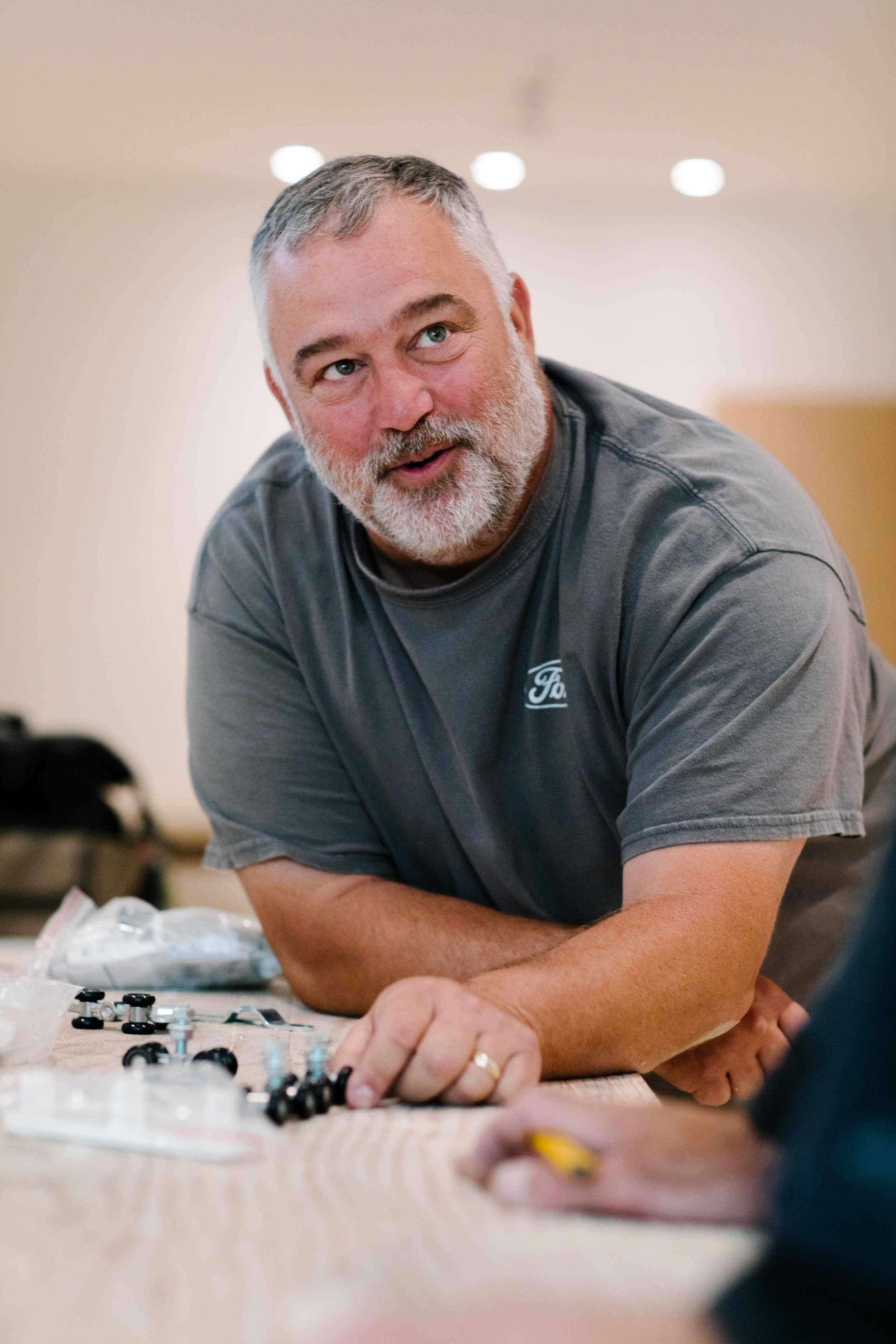 Man with a gray beard leaning on a table, interacting with small hardware pieces.
