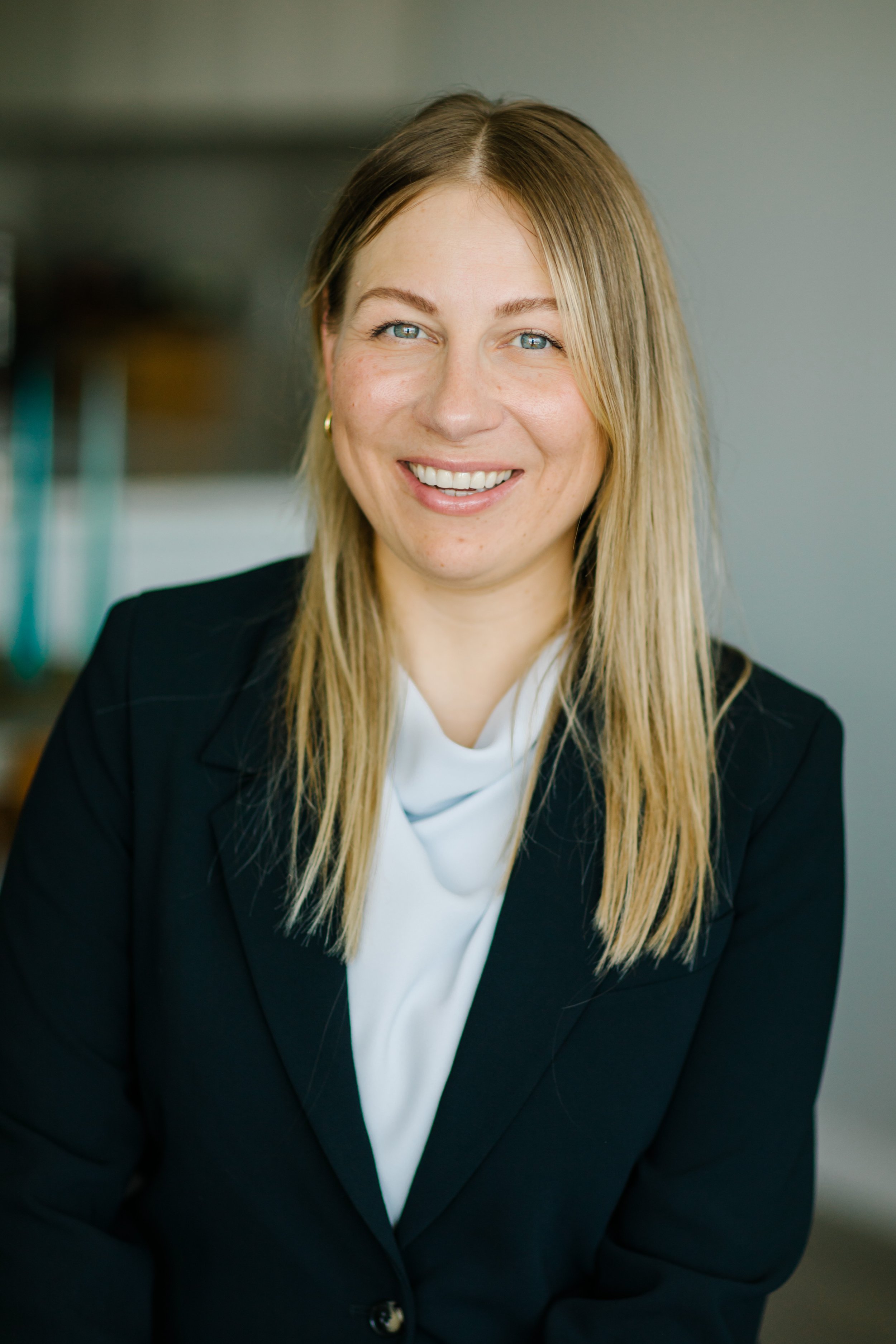 Smiling woman with long blonde hair wearing a black blazer in a neutral background.