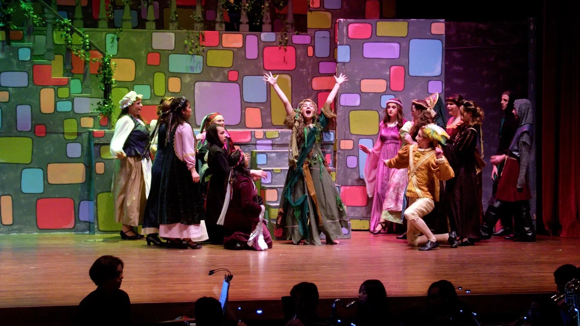 A theatrical performance on stage featuring multiple actors in costumes, with one actor in the center raising their arms. The background is colorful with block-like patterns, indicating a set design for a play or musical.