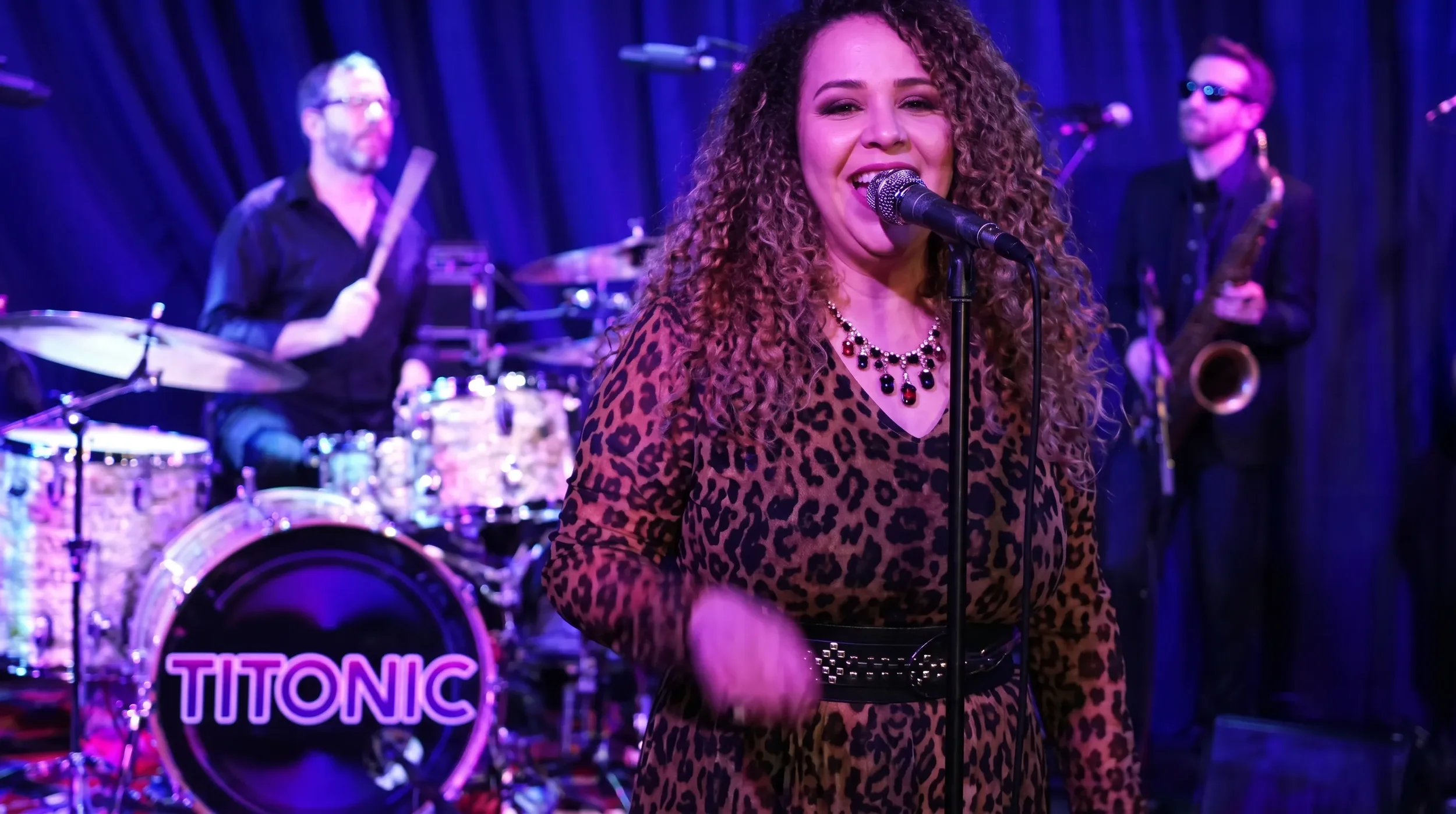 Monique, lead singer for the Colorado band TITONIC stands at microphone with drumer and saxophone player in the background